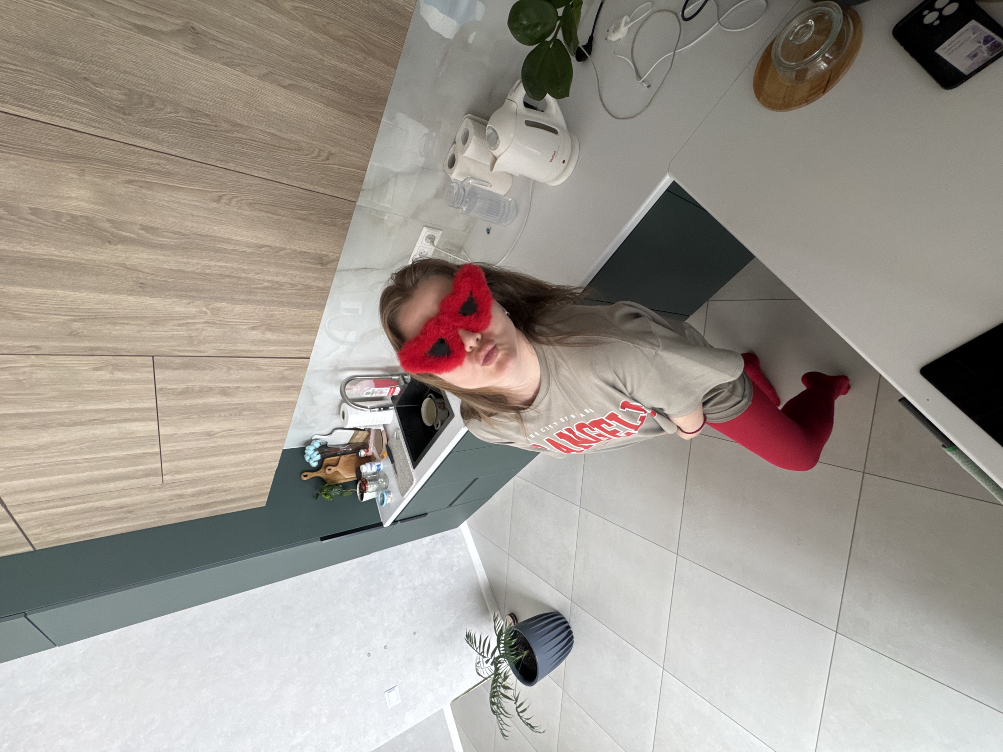 A young woman wearing red fuzzy sleep mask stands in a kitchen in Gornyi, Dvurechenskoe S/P, Russia. She wears a grey t-shirt and red tights. She is standing next to a countertop with a sink and various kitchen items, including a kettle, plant, and cleaning supplies. The kitchen has white tile flooring. A plant in a grey pot is in the corner of the room.