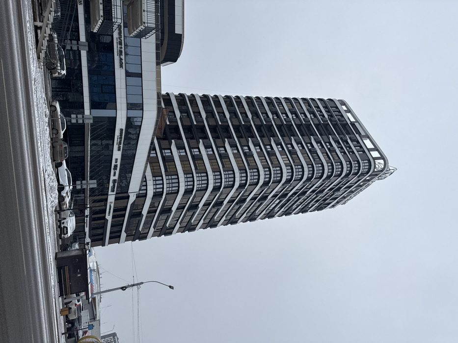 A tall, modern building dominates the scene against a cloudy sky. It features a striking design with curved, white balconies on each floor. Adjacent to it, another building with a glass facade is visible, displaying business names. A street with parked cars and snow lines the foreground. A lamppost stands nearby. The overall impression is urban and somewhat cold, due to the wintery weather.