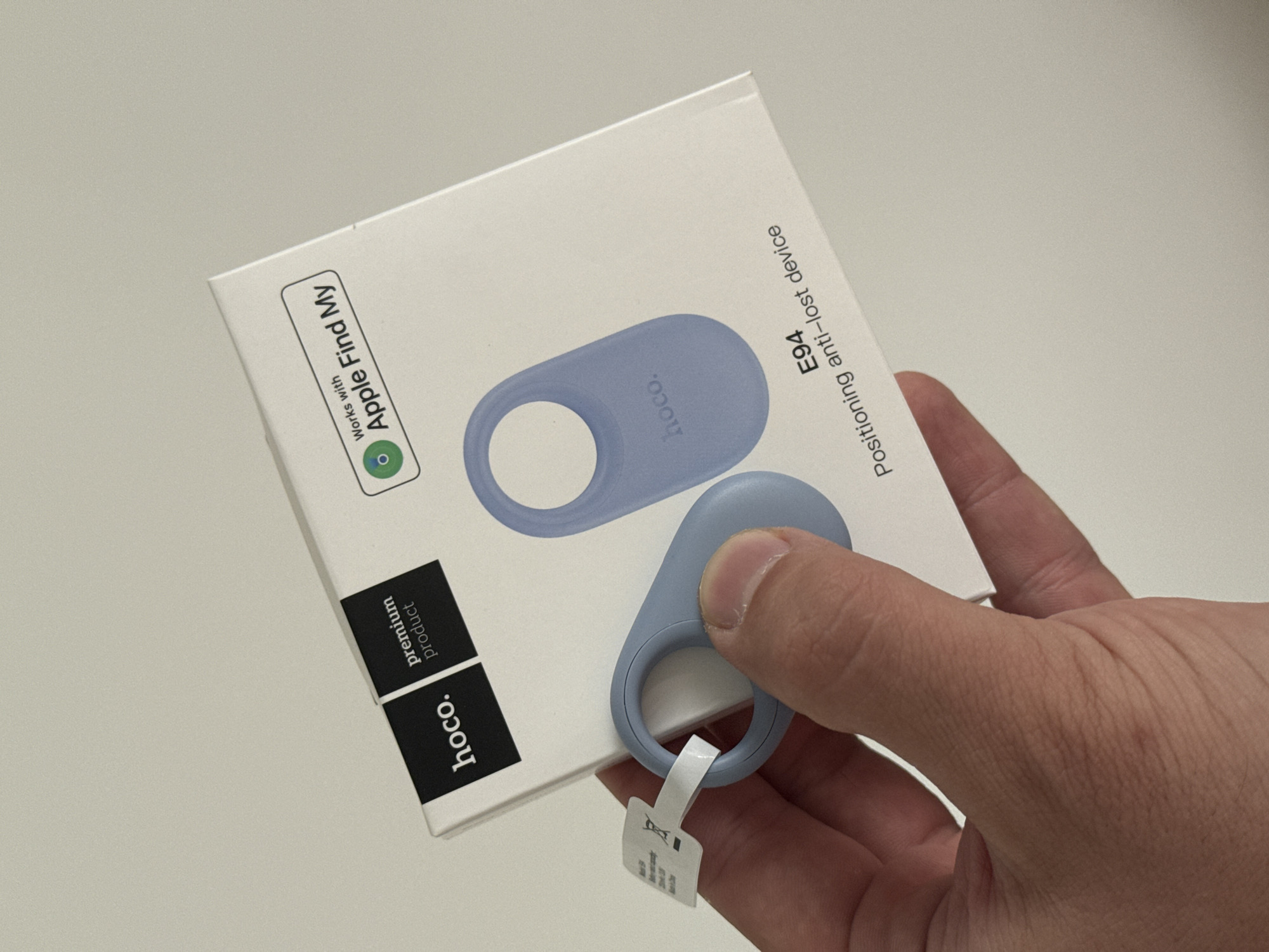 A close-up, high-angle shot in Shirkovo, Russia, presents a human hand holding a white product box and a light blue anti-lost device. The box, branded "hoco." as a "premium product," displays a graphic of the device and features a "Works with Apple Find My" logo. Text on the box identifies the product as "E94 Positioning anti-lost device." The actual light blue device, which is oval-shaped with a circular cutout at one end and "hoco." embossed on its surface, is being held by the thumb and forefinger. A small white pull-tab is partially inserted into the device's circular cutout. The background is a plain, light gray surface.