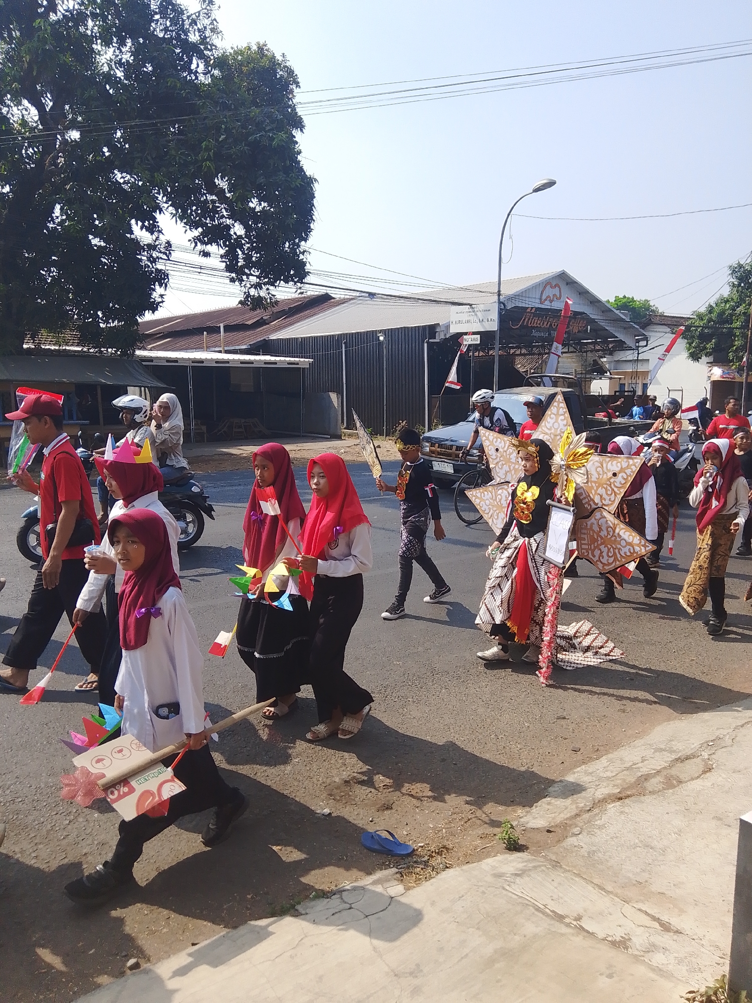 In Kalinyamatan, Indonesia, a parade is in progress on a sunny day. A group of children dressed in festive attire, including headscarves and traditional Indonesian clothing, are walking down a paved road. Some children are carrying small Indonesian flags, while others hold colorful pinwheels. One child is dressed in an elaborate costume resembling a star, adorned with gold and patterned fabric. A few adults, some wearing red shirts and hats, are also participating in the procession. A motorcycle is visible in the background, and a few buildings line the street, one with a sign that reads "Maestro." The environment appears to be a semi-urban area with trees and overhead power lines contributing to the scene.
