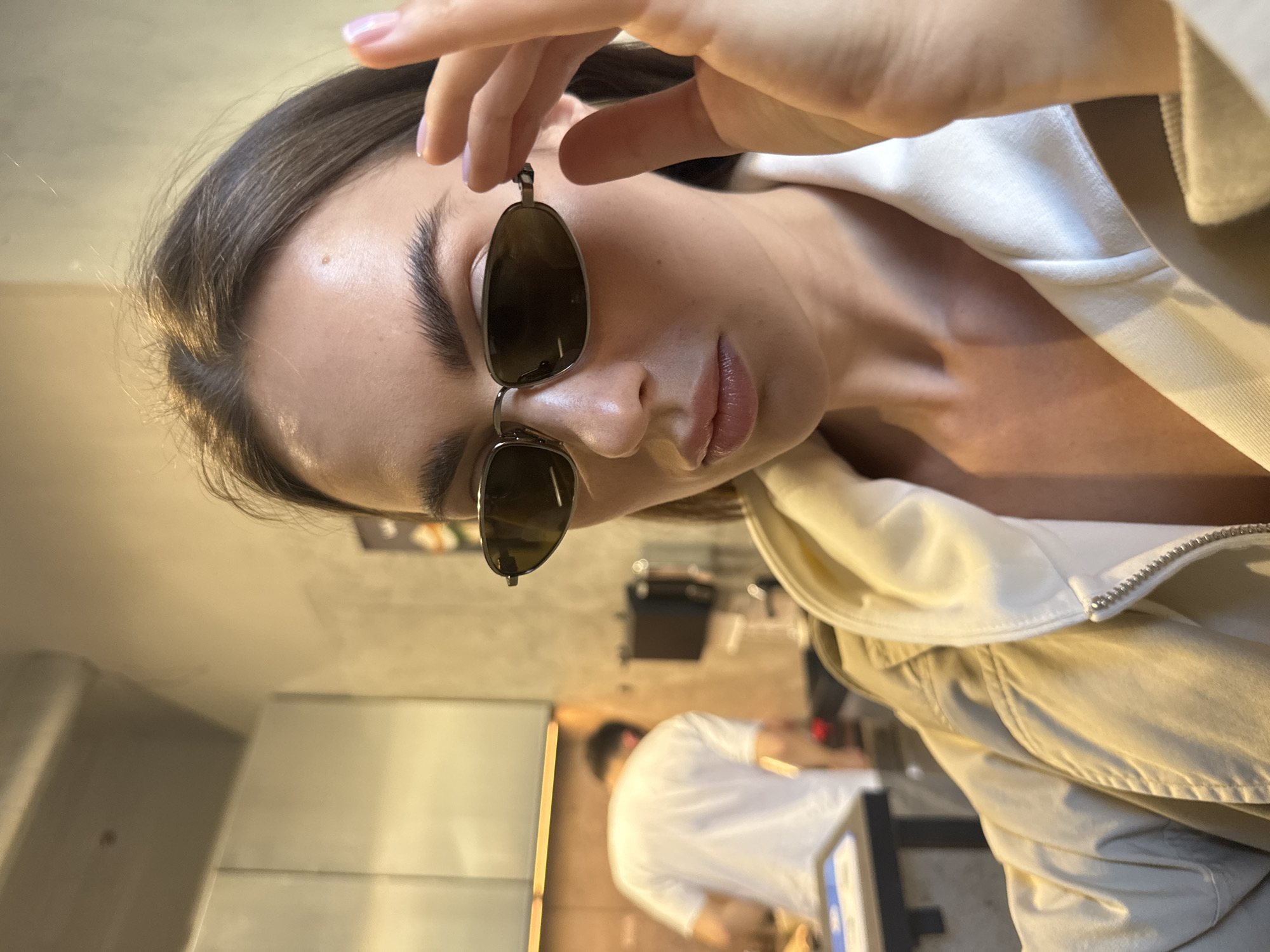 The image shows a young woman posing indoors, captured from a tilted, slightly low-angle perspective. She has dark hair, prominent eyebrows, and is wearing a light-colored top beneath a beige jacket. Her right hand, with light pink painted nails, holds a pair of dark-lensed sunglasses with silver frames near her face, partially covering her eye. Her lips are slightly parted, giving her a subtle, composed expression.
In the blurred background, another person wearing a white t-shirt is visible, bent over an unseen object, suggesting they are engaged in an activity like working or preparing something. The setting appears to be a modern indoor space, possibly a cafe, restaurant, or a contemporary kitchen in Warsaw, Poland. A stainless steel appliance, perhaps a refrigerator or counter, is visible on the left side of the frame. The lighting is artificial, characteristic of an interior environment. There is no visible text in the image.