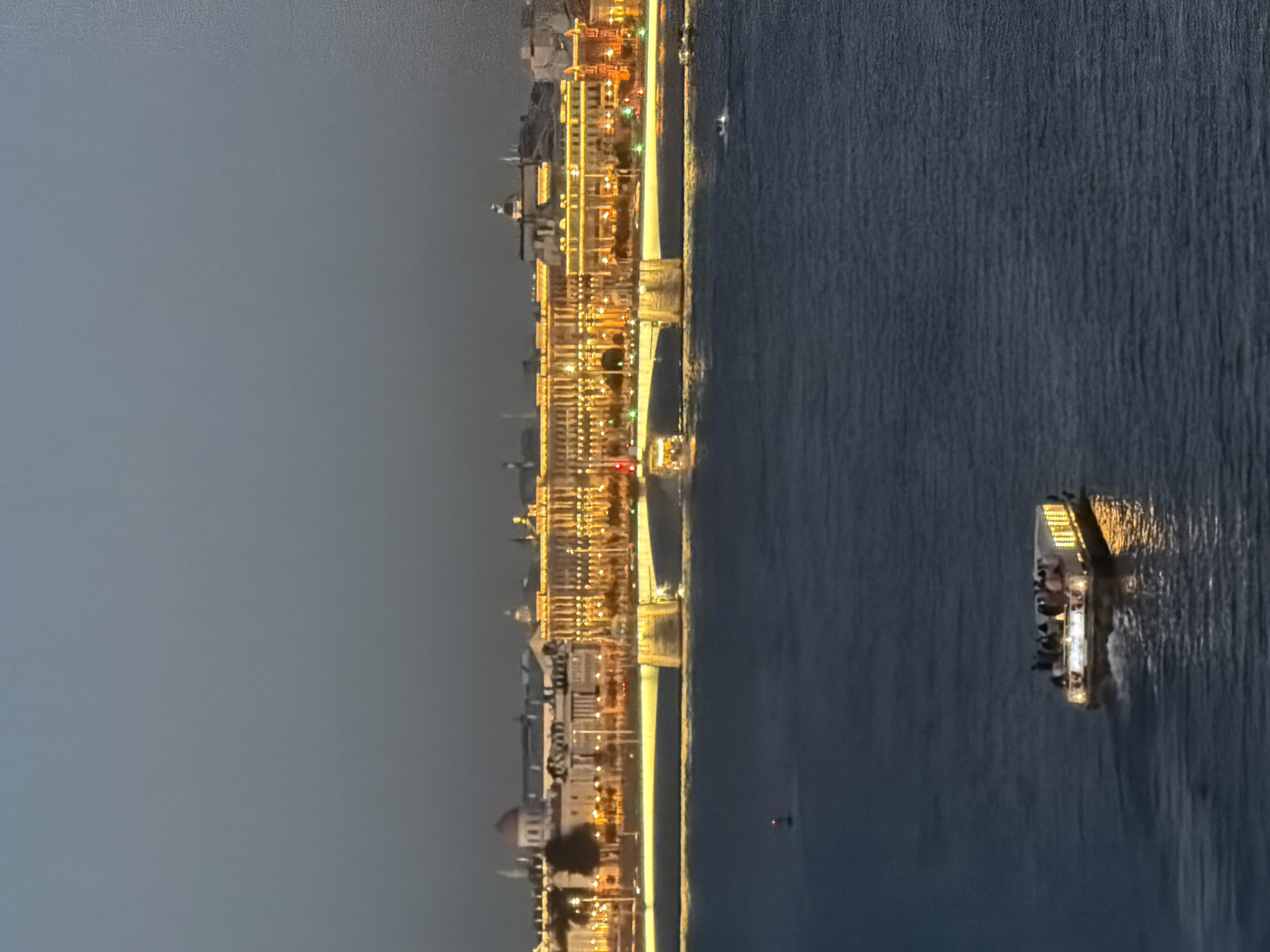 The image shows a nighttime view of the city of Saint Petersburg, Russia, from across a body of water, likely the Neva River. In the background, a long, illuminated building with numerous windows and decorative lights stretches across the horizon. A bridge, also lit with yellow lights, spans the water in front of the building. In the foreground, a small boat filled with people is moving across the dark water, its interior lights casting a warm glow. The sky is a dark, twilight blue, and the water is a deep, dark blue, reflecting some of the lights from the city. A single red navigational buoy is visible in the water to the left of the boat. The overall scene depicts a peaceful evening in a historic city.