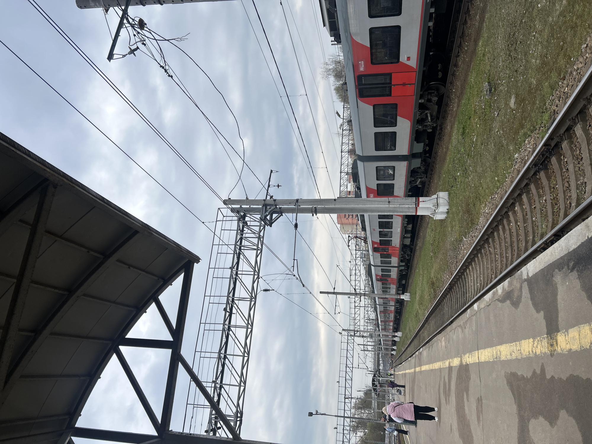 This photograph captures a railway station scene in Klin, Russia, under an overcast sky. The image shows a long station platform on the right, made of concrete or asphalt with a distinct yellow safety line running its length. The platform surface appears somewhat damp or stained. Several people are visible on the platform, with one individual in a light pink jacket and dark trousers standing more prominently in the mid-ground, facing away from the camera.
Parallel to the platform, two sets of railway tracks extend into the distance. A modern commuter train, featuring a red and grey/white livery, is stopped on the tracks further from the platform. The acronym "ЦППК" (Central Suburban Passenger Company) is clearly visible in red letters on the train's side. A strip of green grass separates the train from the tracks closer to the platform.
Above the tracks and platform, a dense network of overhead power lines and robust metal support structures, including poles and gantries, stretches across the frame, indicating an electrified railway system. A section of a dark metal-framed station canopy or roof is visible in the upper left corner of the image, partially obscuring the cloudy sky. The overall lighting suggests an overcast day, without direct sunlight.