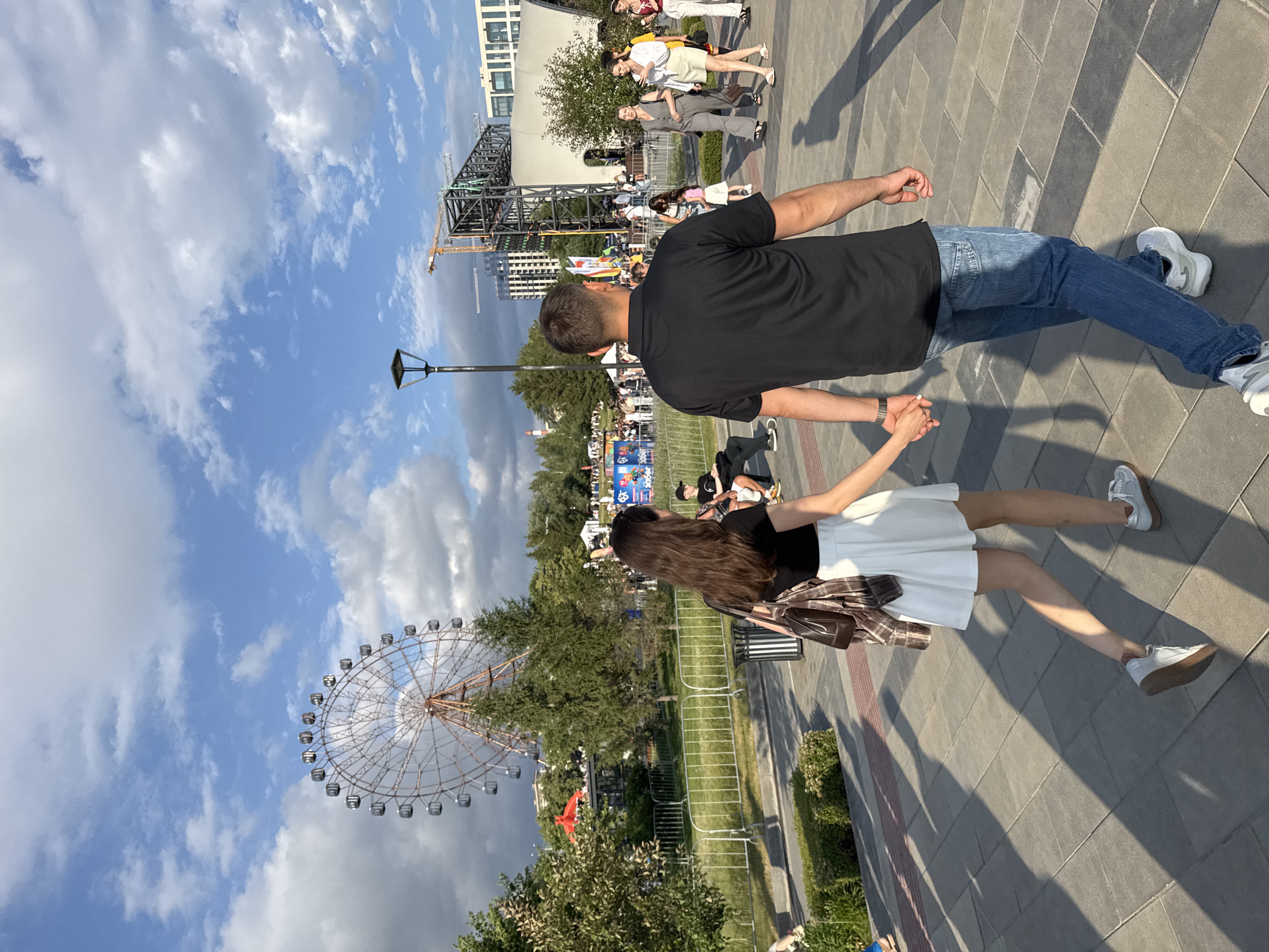 A couple walks hand-in-hand on a paved path in a park with a ferris wheel in the background. The woman is wearing a black crop top and a white skirt, while the man is wearing a black polo shirt and blue jeans. They are walking away from the camera. In the background, there is a ferris wheel, trees, buildings under construction, and other people walking around. The sky is blue with some clouds. The location is identified as Novosibirsk, Russia.