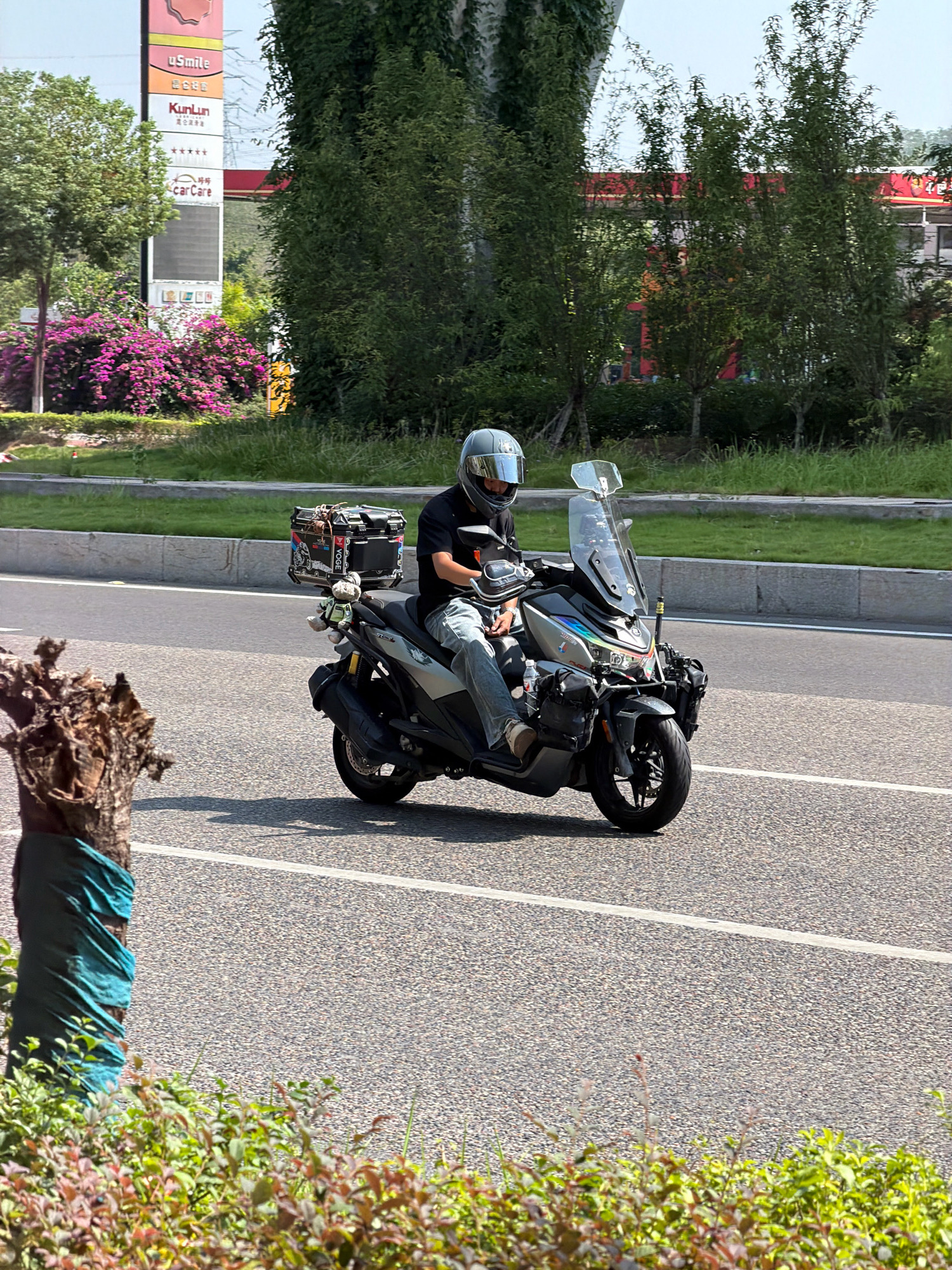 A person wearing a silver helmet and dark clothing is riding a silver scooter on a paved road. The scooter has a large black storage box mounted on the rear and other accessories attached to the front and sides, including a small plush toy hanging from the back. The rider is wearing blue jeans and tan shoes. The road is bordered by a grassy area on the left and a sidewalk with trees and a billboard on the right. The billboard displays "uSmile" and "KunLun". In the background, there are buildings and lush green trees. The image appears to have been taken during the daytime under a clear sky. The presence of the KunLun billboard suggests the location is likely in China, possibly in Jiangjin, a district of Chongqing, China, as it is a common brand found in that region.