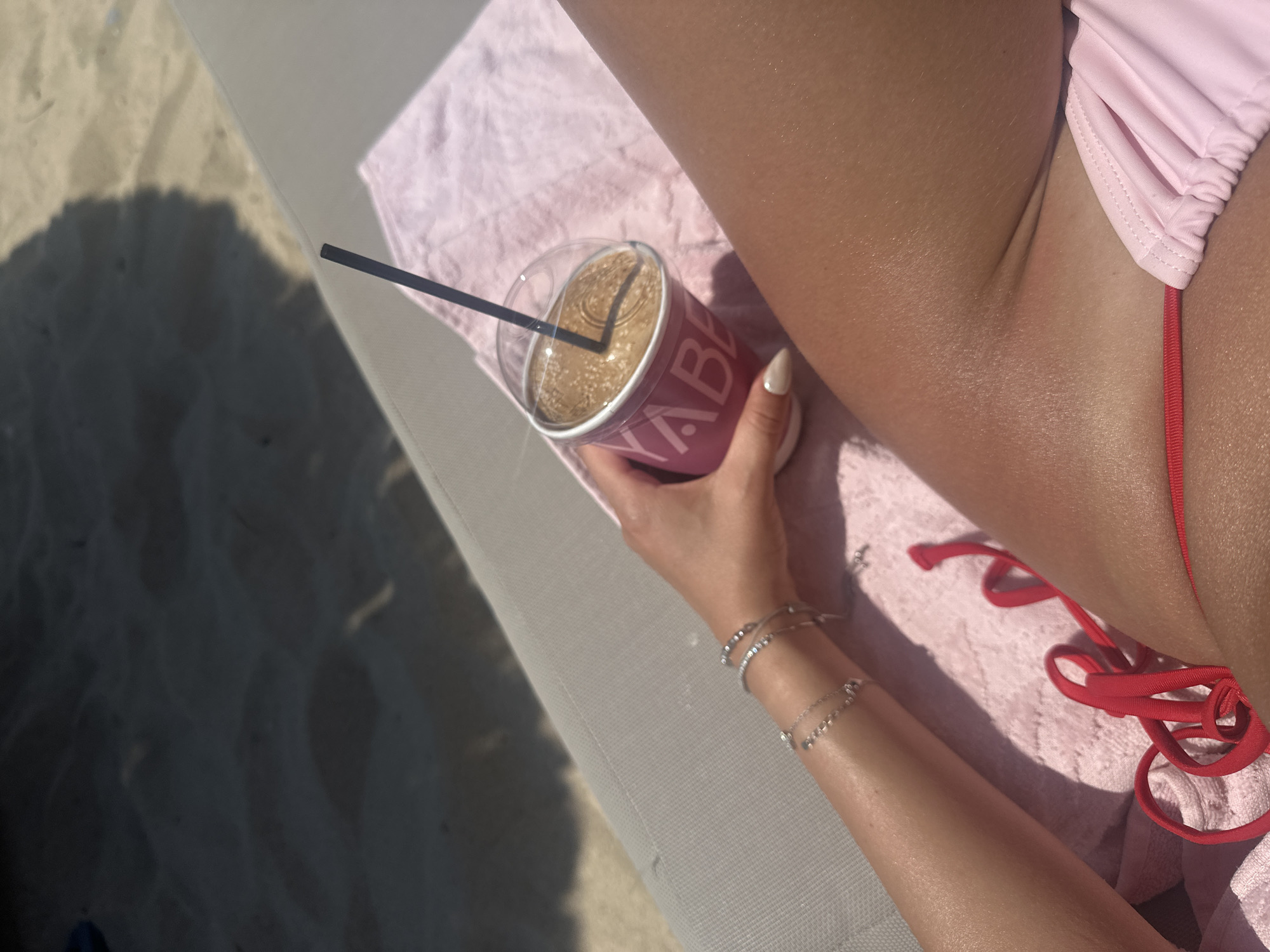 The image shows a close-up of a person's arm and leg resting on a pink towel, on what appears to be a beach lounger. The person is holding a translucent cup with a dark straw, containing a frothy brown beverage. The cup has pink lettering that partially reads "YABER". The person is wearing a pink bikini bottom with red ties and several silver bracelets on their left wrist. The background shows sand and the shadow of an umbrella. The scene is lit by sunlight, suggesting it is daytime. The location is Ofrynio Beach, Greece.
