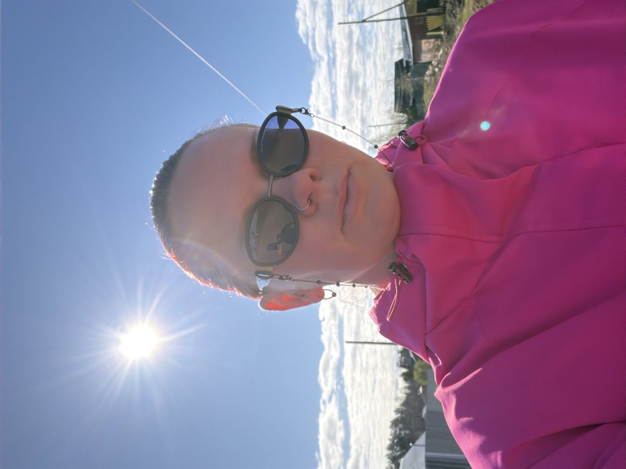 A person wearing sunglasses and a bright pink hooded jacket is the primary subject, facing towards the camera. The person has a nose ring. The sky is blue with the sun prominently visible. Trees and a building are visible in the background, along with puffy white clouds. It is unclear where in Chaikovsky, Russia this photo was taken.
