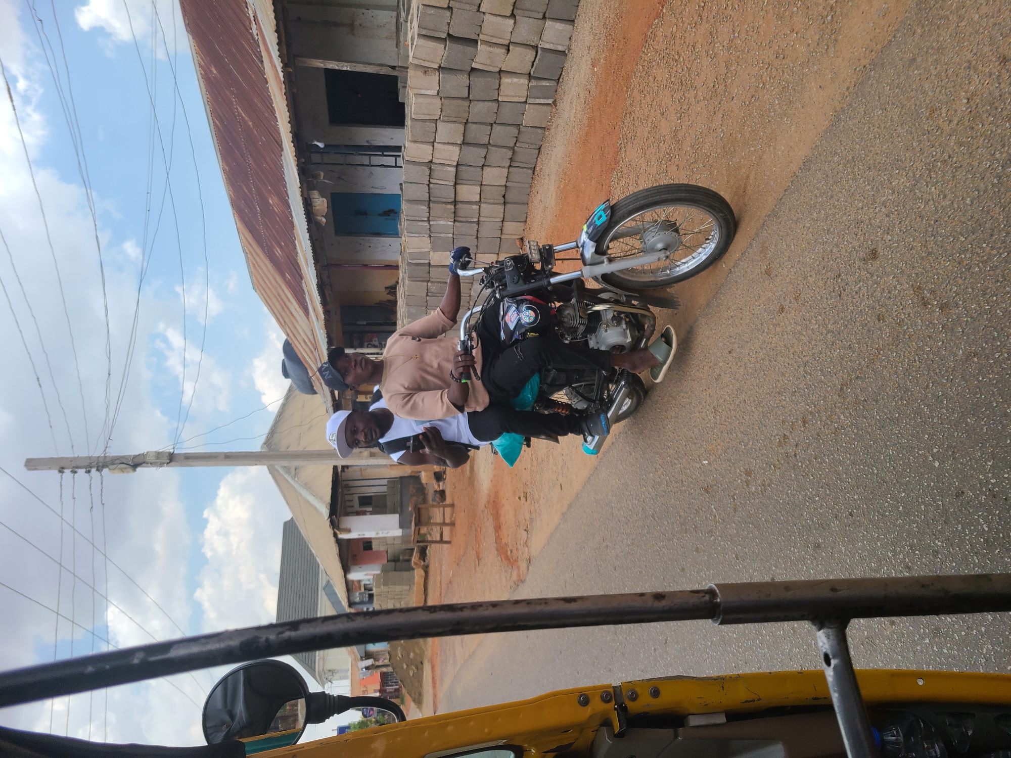 Two men are riding a motorcycle on a paved road in Jos, Nigeria. The driver is wearing a tan shirt, black pants, a dark-colored cap, and gloves. The passenger is wearing a white shirt, black pants, and a white cap. The motorcycle is black and silver. The background includes buildings constructed of cinder blocks and corrugated metal, a utility pole with wires, and a dirt shoulder on the side of the road. A yellow motorized tricycle is partially visible in the foreground. The sky is partly cloudy.