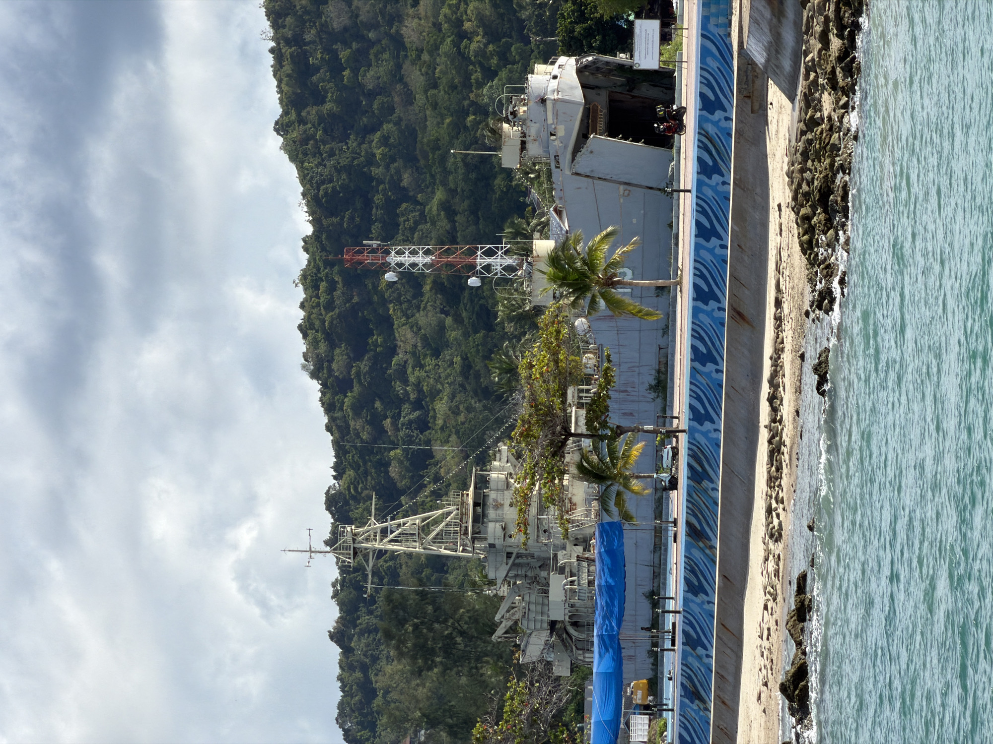 This image depicts a waterfront scene in Koh Phangan, Thailand. The primary subjects are two docked ships, one appearing larger and more derelict than the other, suggesting they might be decommissioned or undergoing renovation. In the background, a lush, green, tree-covered hill rises steeply. A red and white communication tower stands tall near the first ship, with a palm tree in front of it. The foreground shows a stone-lined seawall with a blue, wave-like patterned design, bordering a beach area with scattered rocks and then the clear, turquoise ocean.
The scene appears to be set during the daytime, with bright but partly cloudy skies. The presence of the communication tower and the ships suggests a possible port or a historical site related to maritime activities. There is no visible activity or event taking place, giving the image a static and somewhat tranquil feel, despite the potentially industrial nature of the ships. The water is calm, with gentle waves lapping against the shore. The overall impression is one of a coastal landscape that blends natural beauty with man-made structures. No discernible text is visible in the image.