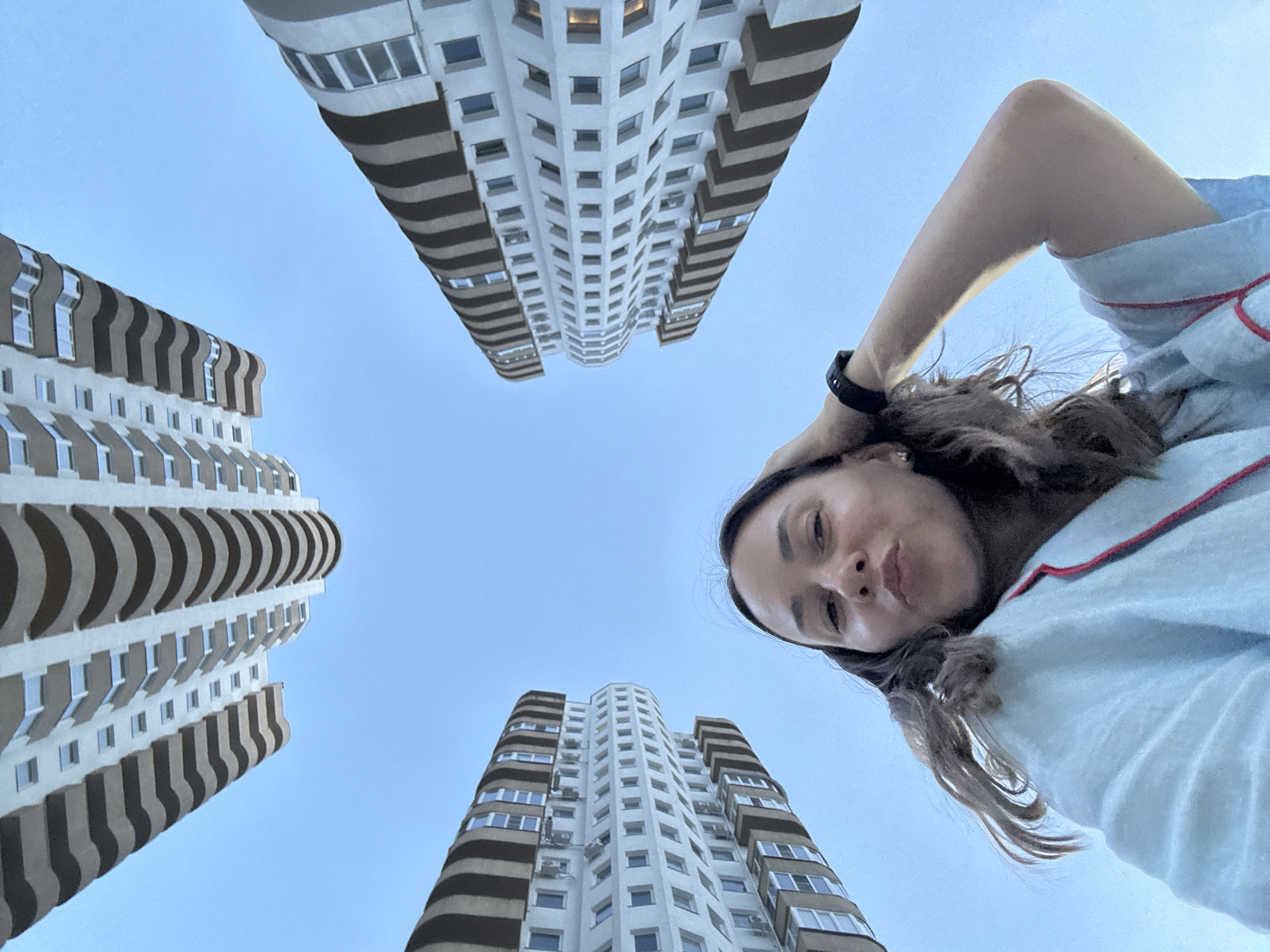 In Sochi, Russia, a young woman with dark, wavy hair is captured in a low-angle, fisheye perspective selfie. She is wearing a light blue robe with red trim and a black smartwatch on her left wrist. Her right hand is casually resting on her head, and she is puckering her lips towards the camera. The background is dominated by the clear blue sky, and surrounding her are the curved facades of modern apartment buildings, their repeating balconies creating a visually striking pattern. The buildings appear to be constructed with white and dark brown elements. The overall impression is of a relaxed moment in an urban setting.