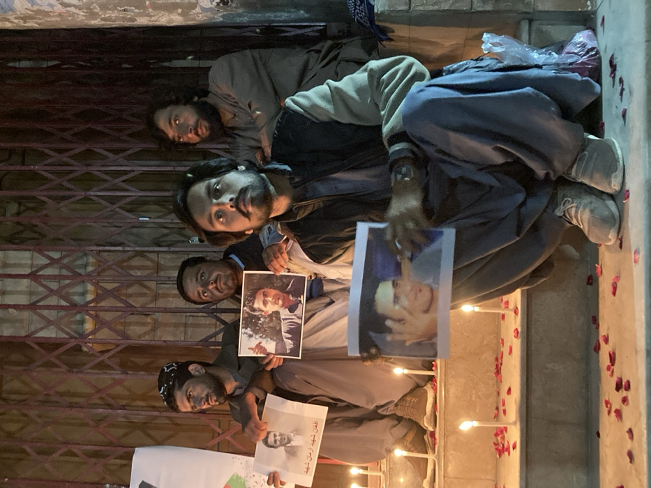 Four males are depicted at night in Quetta City Tehsil, Pakistan, participating in what appears to be a candlelight vigil. They are seated on stone steps against a dark, collapsible metal security gate. The foreground features numerous lit white pillar candles and scattered red rose petals.
One male, positioned in the lower right, squats and holds a horizontal photograph of a smiling man. He wears a dark vest over a light-colored tunic, dark loose trousers, and light-colored athletic shoes. A watch is visible on his left wrist. Adjacent to him, a transparent plastic bag containing rose petals rests on the step.
Behind him, a second male sits on a higher step, holding a horizontal photograph of a man giving a thumbs-up gesture. To the left, a third male sits on the same step, wearing a patterned skullcap and light-colored attire, holding a vertical photograph. This photograph displays a man's portrait with Urdu text below it, translating to "Afghan Shaheed Arman Shaheed."
A fourth male is partially visible in the upper right background, seated on a higher step, wearing a light-colored tunic. All individuals appear to be focused, with somber expressions, under the illumination of the candles. A partially visible white poster with red and green elements is on the far left, and a blue fabric item is on the far right.