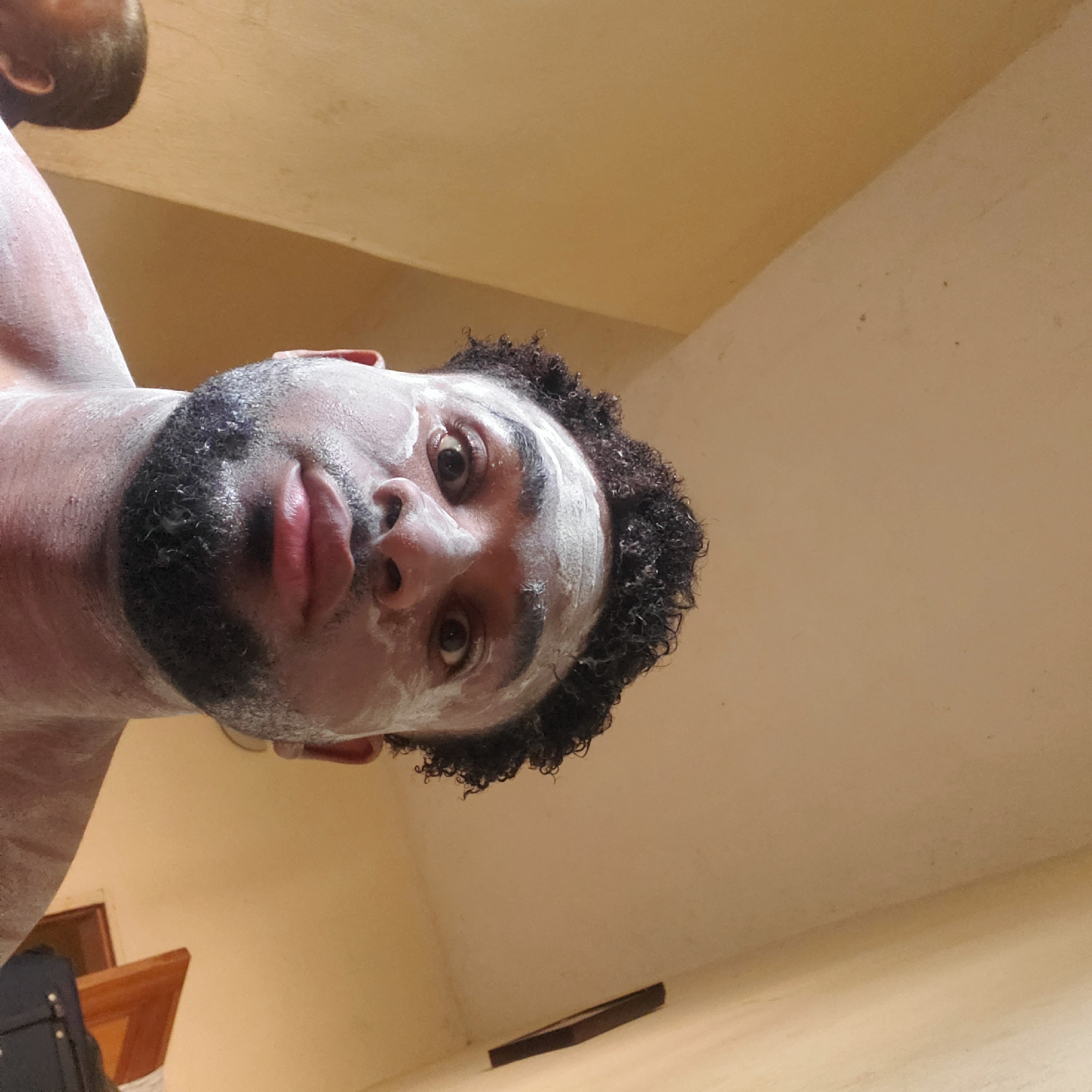 A low-angle, close-up shot depicts a man with dark curly hair and a beard. His face and upper chest have a white substance applied, possibly a cream or mask. He is looking down at the camera with a neutral expression. In the top left corner, a partial view of another person's head is visible. The background consists of a cream-colored wall and ceiling. Other objects are partially visible, including a brown frame and a black bag. There is no indication in the image that the location is Obosi, Nigeria.