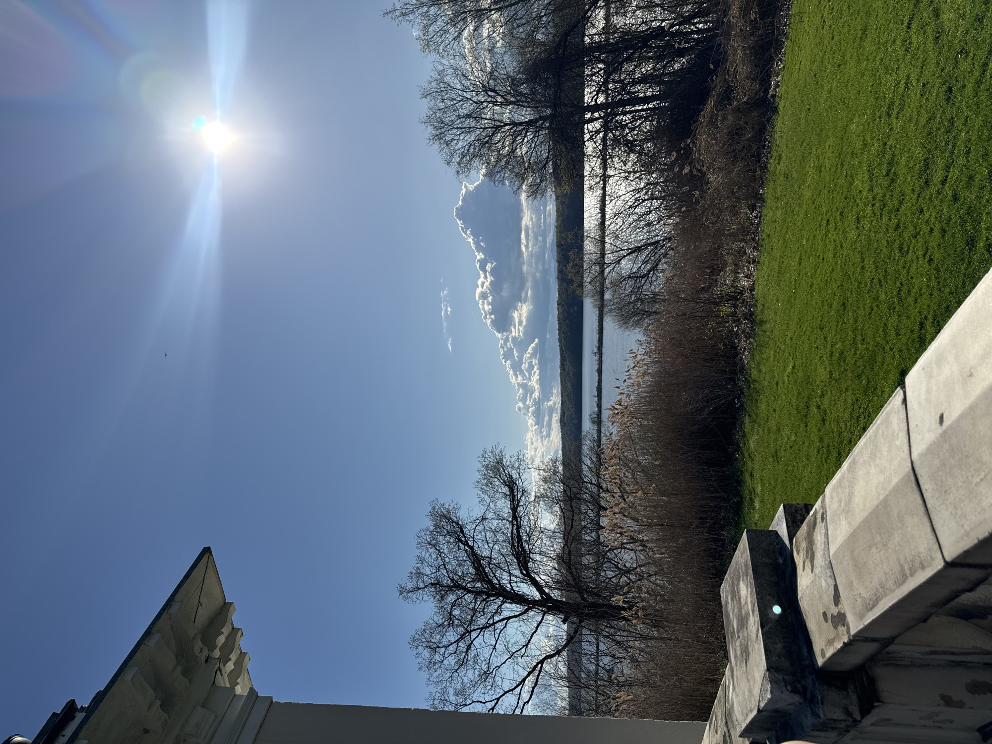 The image presents a sunlit outdoor scene in the Town of Red Hook, United States of America. The viewpoint is elevated, likely from a structure with concrete walls visible in the lower right and a peaked roof on the lower left. The upper left is dominated by a bright sun in a clear blue sky, which causes a lens flare effect. The sun’s glare is intense.
A narrow body of water extends horizontally across the center of the frame, possibly a lake or wide river. The water reflects the sky and some clouds. Fluffy white clouds are visible in the sky above the water, adding depth to the scene. The far bank of the water is lined with trees, mostly bare of leaves, and some tall reeds or grasses in the foreground.
To the right of the trees, a patch of green grass is present, suggesting a field or lawn. The lighting indicates it is daytime and likely during the late fall or winter season, considering the bare trees. There are no people visible in the frame.
