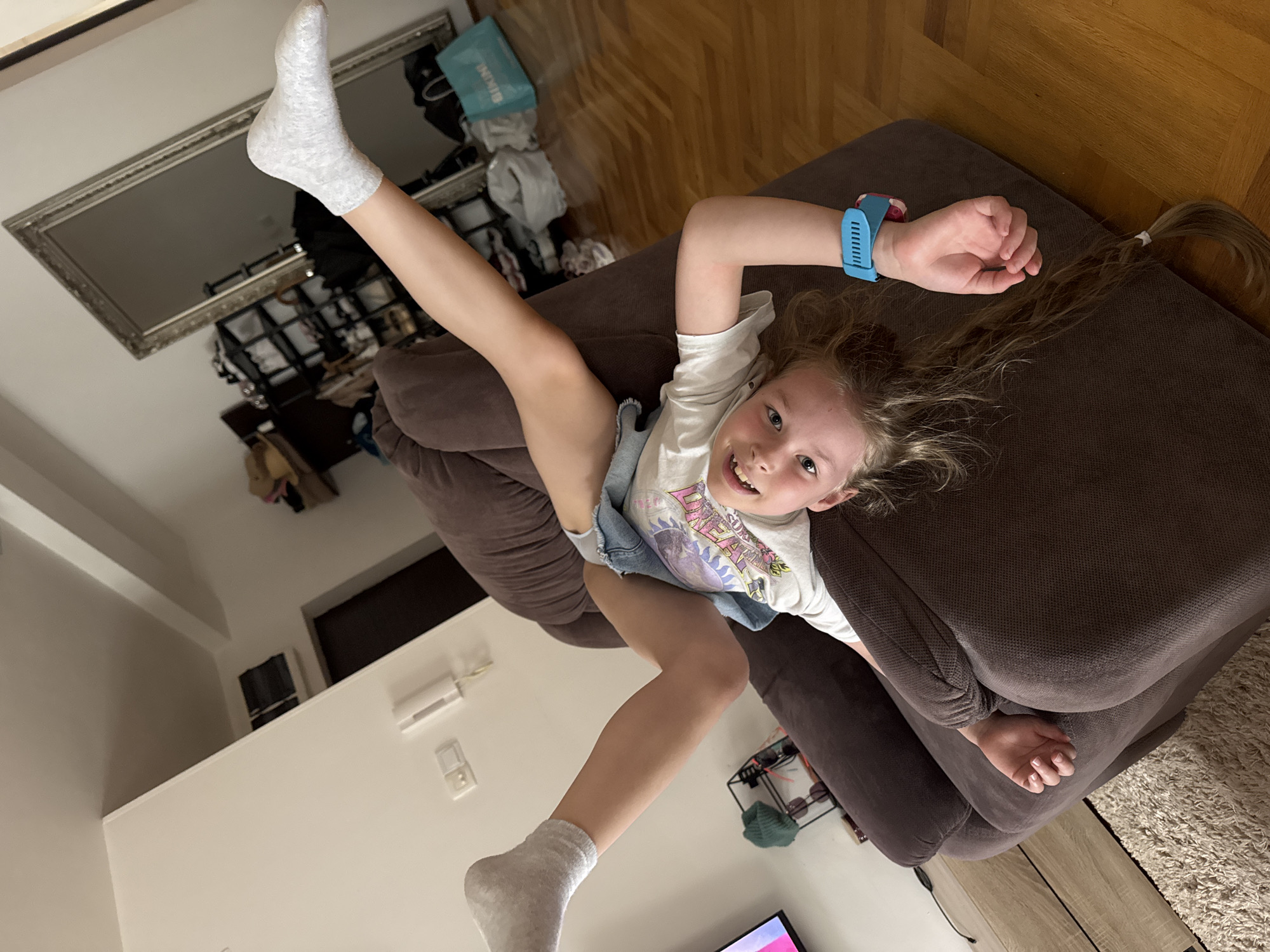 A young girl is pictured in a living room. She is lying on a dark brown couch, with her head near the right side of the frame and her legs raised high on the left. She is wearing a white t-shirt, a denim skirt, and grey socks. A blue smartwatch is visible on her left wrist. Her hair is partially tied back in a ponytail.
Behind her, there is a silver-framed mirror on the wall, reflecting cluttered shelves. The room has a wooden floor and white walls. A television and other household items are visible in the background.
There is no evidence to determine if this picture was taken in Budva, Montenegro.