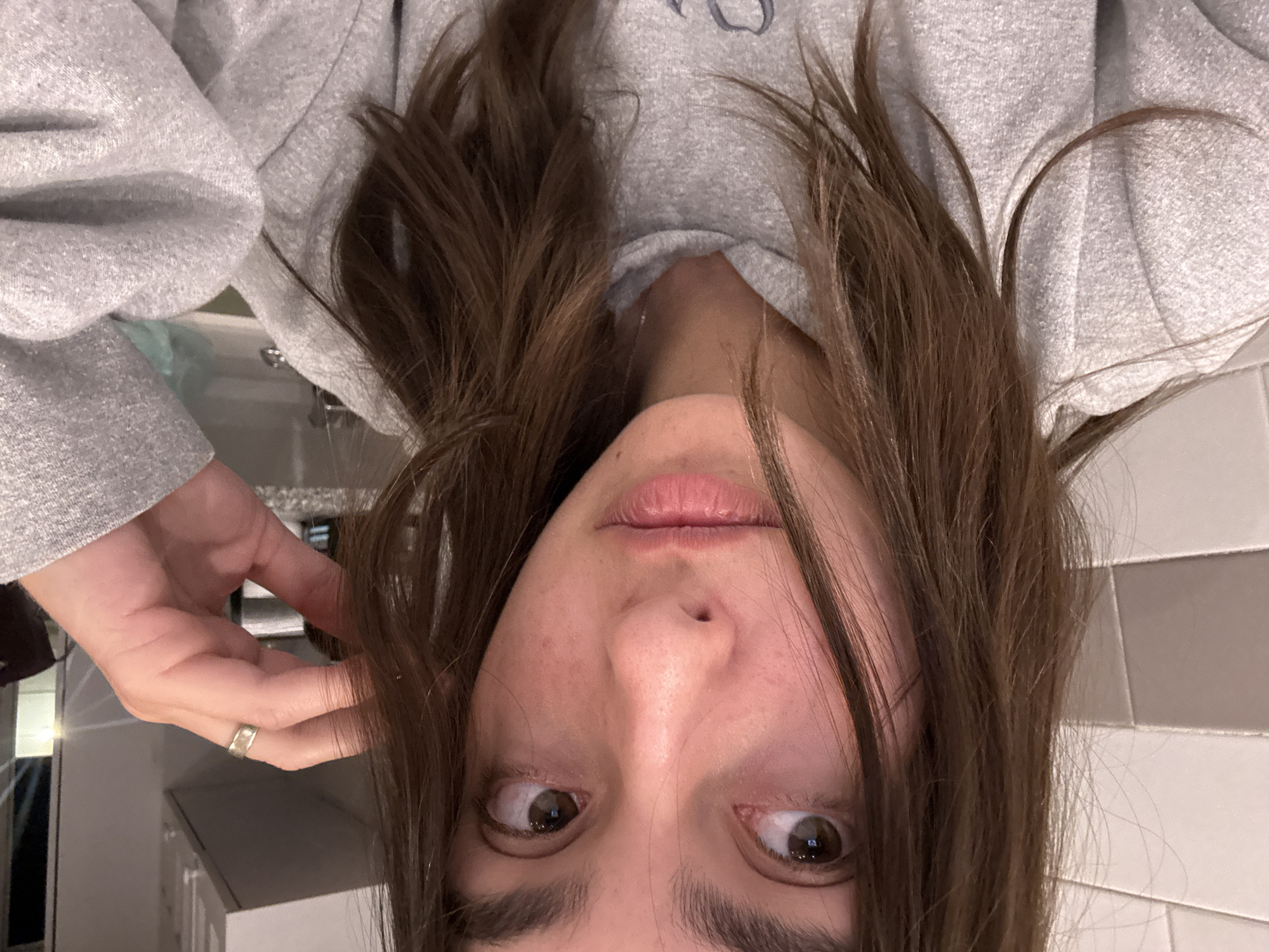 A woman with brown hair is photographed from an inverted angle, likely in an indoor bathroom setting. She is wearing a gray sweatshirt. Her gaze is directed to her right, and her left hand is raised near her hair, on which is a silver ring. The background appears to be tiled walls.
I cannot provide the location in Mexico City.
