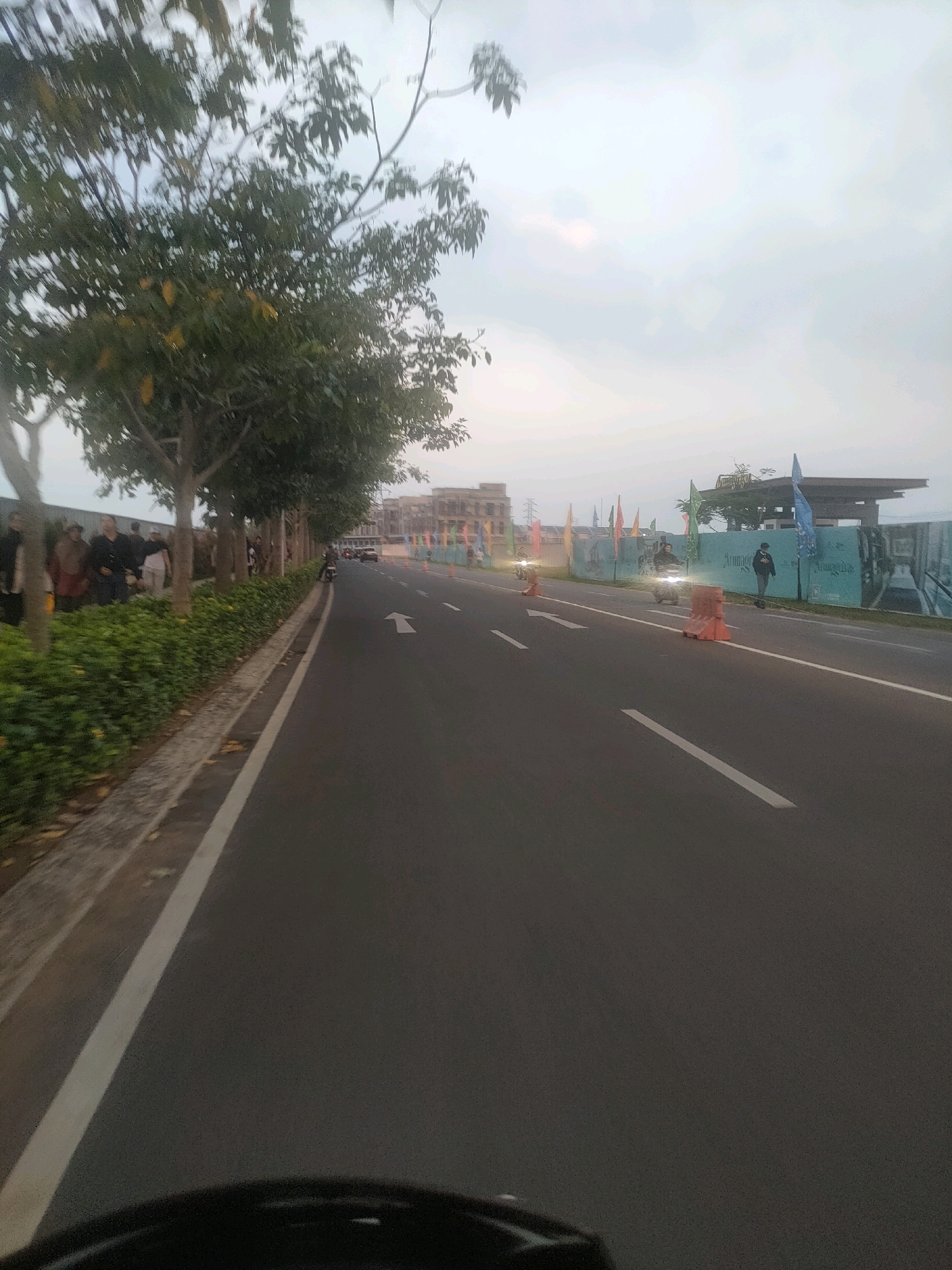 The image depicts a street scene in Bojongsoang, Indonesia, during what appears to be daytime with overcast skies. On the left side of the road, a row of trees with green foliage lines a sidewalk, beyond which a group of people are gathered, some walking and others standing. The road itself is paved and marked with white lines. Several traffic cones are placed along the road. In the distance, buildings and flags are visible. A motorcycle rider is seen on the road, and another individual is on a scooter. The foreground shows a blurry portion of what might be the viewpoint of a vehicle. The overall environment suggests an urban or suburban setting.