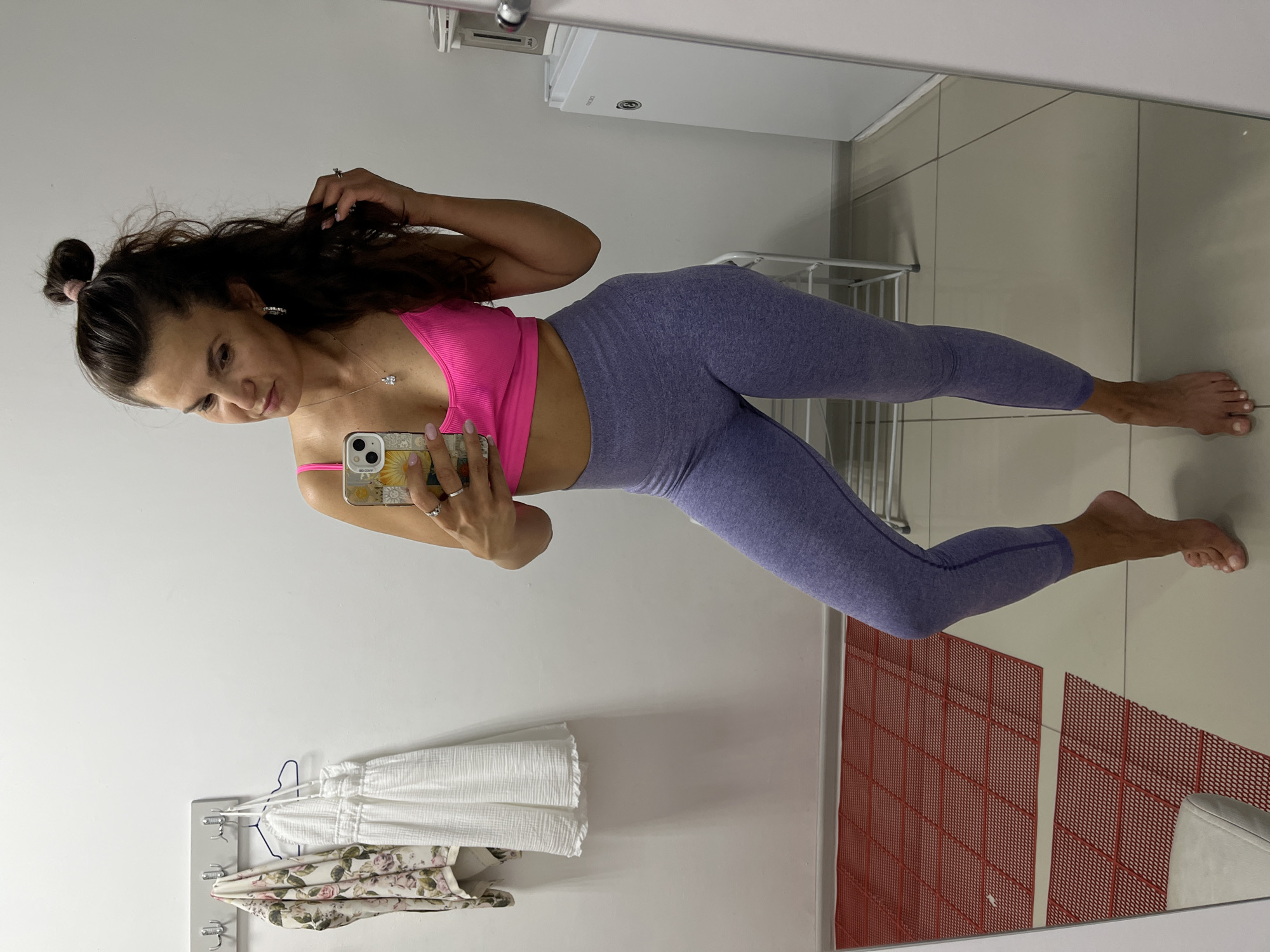 In Petropavlovsk-Kamchatsky, Russia, a woman stands in what appears to be a dressing room or a small personal space. She is wearing a bright pink sports bra and lavender-colored leggings. The woman is holding a smartphone in her right hand, taking a mirror selfie. Her left hand is casually touching her hair, which is partially pulled up into a messy bun. She is wearing rings on her fingers and small hoop earrings.
The background is dominated by a white wall with a set of hooks holding two garments: a floral-patterned cloth and a white, textured dress. A large mirror reflects the woman and part of the room. The floor is tiled, with a red, grid-patterned mat visible in the reflection. To the right of the woman, a white appliance, possibly a small refrigerator or cooler, is partially visible. The lighting is neutral and diffused, suggesting an indoor setting.
