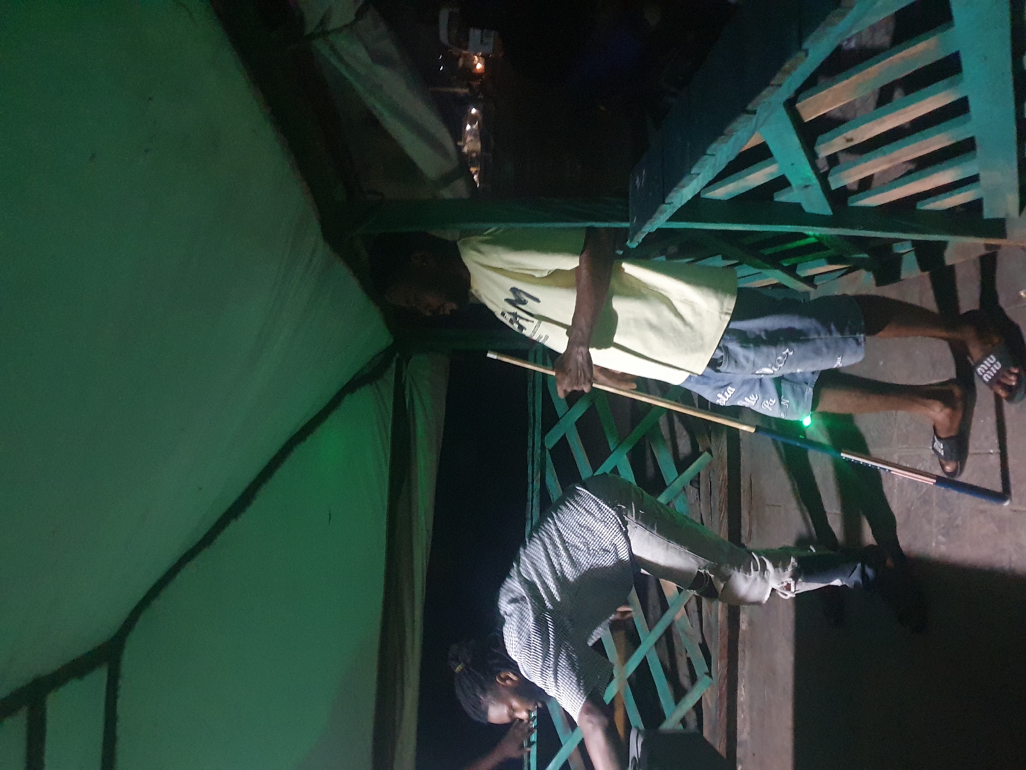 Two men are playing billiards at night under a green tent structure. The man on the left, with dreadlocks, is wearing a checkered shirt and ripped jeans, bending over the pool table. The man on the right, wearing a yellow t-shirt and denim shorts, is holding a cue stick. In the background, a white vehicle and some lights are visible. The overall setting appears to be an outdoor recreational area, possibly in the city of Lokoja, Nigeria, indicated by the lighting and the presence of a vehicle typical of the region. The green tent provides shelter, and the green railing suggests a raised platform or balcony.