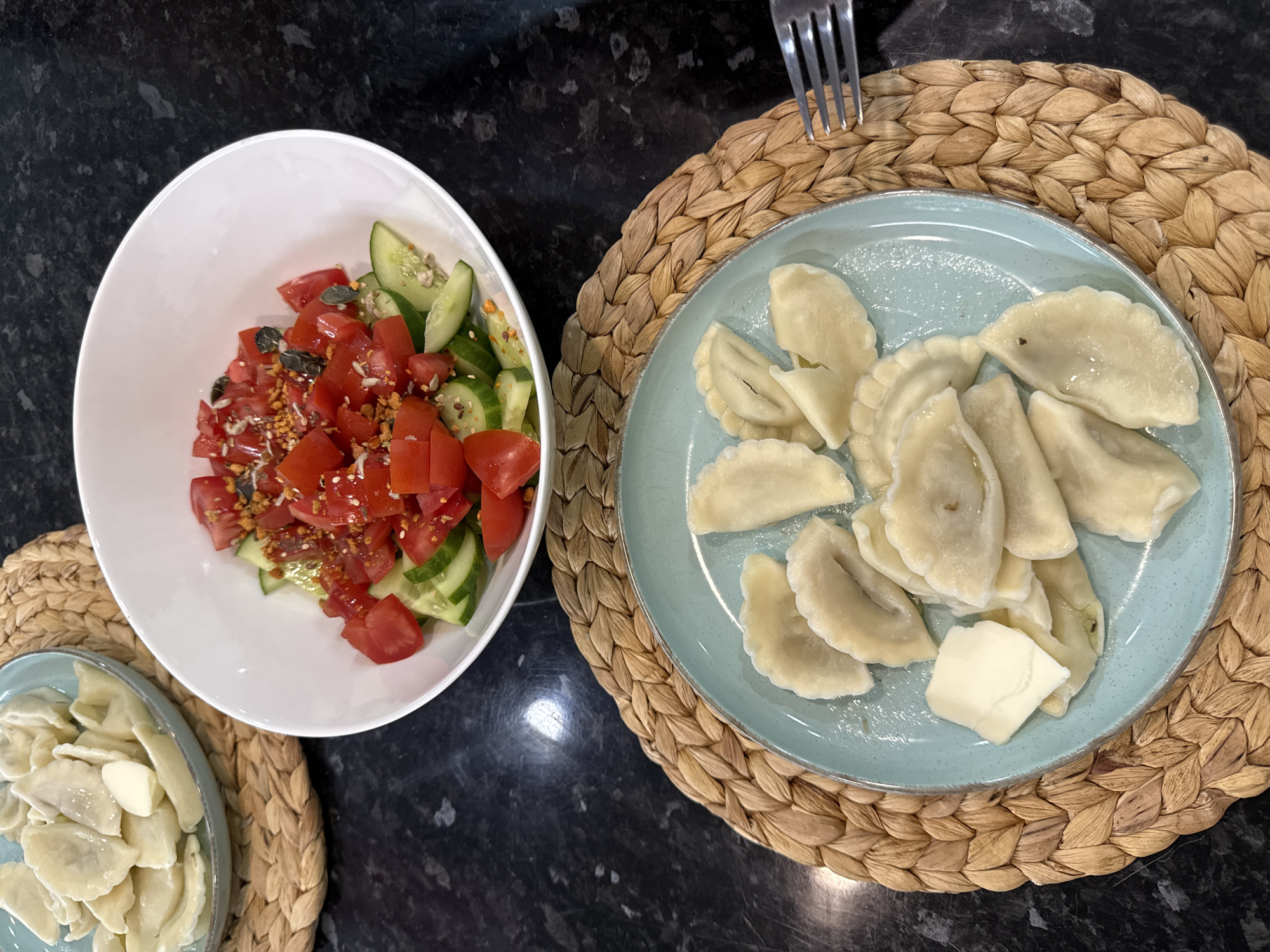 A top-down shot presents two dishes on a dark countertop. A white bowl holds a salad of chopped tomatoes and cucumbers, sprinkled with seeds. To the right, a light blue plate is topped with pierogi and a square of butter, with a fork resting above. Both plates are set on straw placemats.