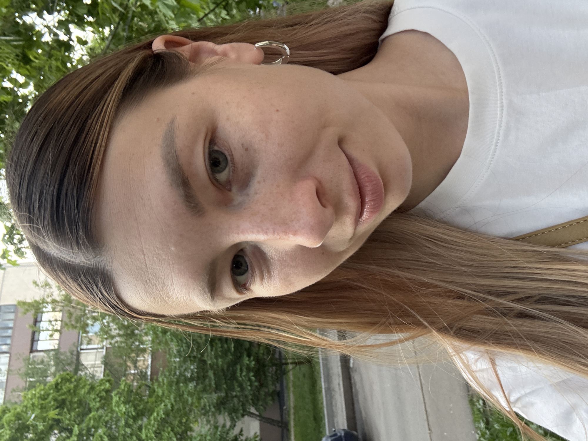 A fair-skinned woman with light brown hair is centered in a selfie-style shot. She is wearing a white t-shirt and silver hoop earrings. Her facial features are prominent, including her eyes, nose, and lips. The background shows green trees and a building, suggesting an urban setting. The location is identifiable as Moscow, Russia based on the building's architectural style, which is typical of Soviet-era construction.