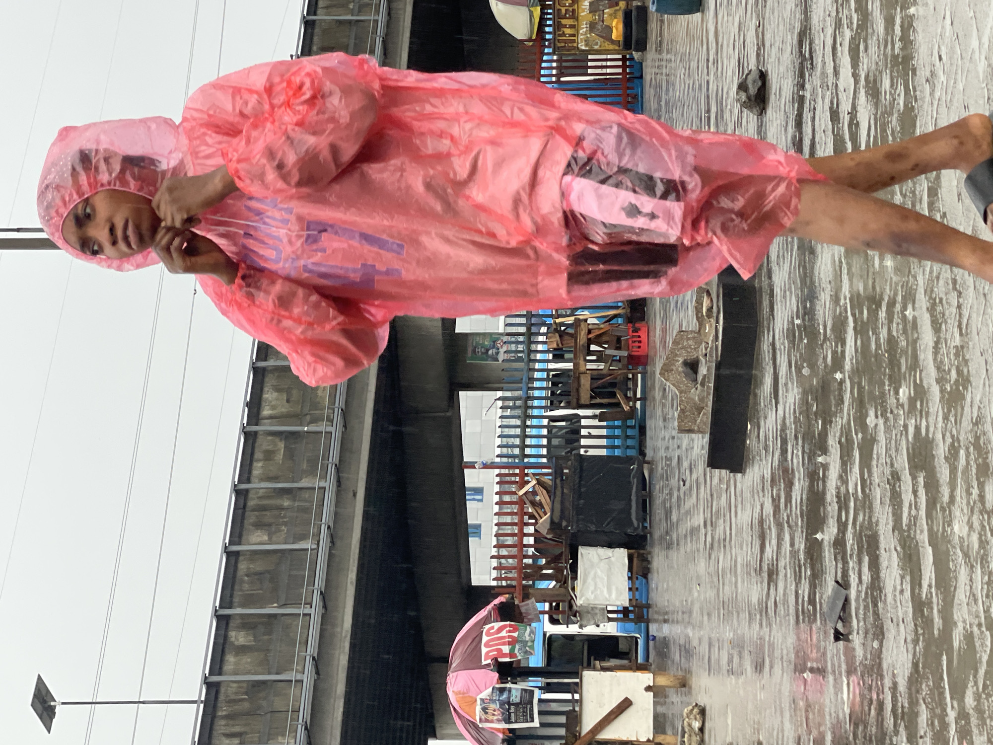 A person wearing a pink plastic raincoat stands in the rain on a flooded street in Lekki, Nigeria. The person has bare legs and is adjusting the raincoat. In the background, there are structures that appear to be part of an elevated road or bridge. There are also various objects like wood and metal pieces. The scene is overcast and wet, indicating recent or ongoing rainfall.