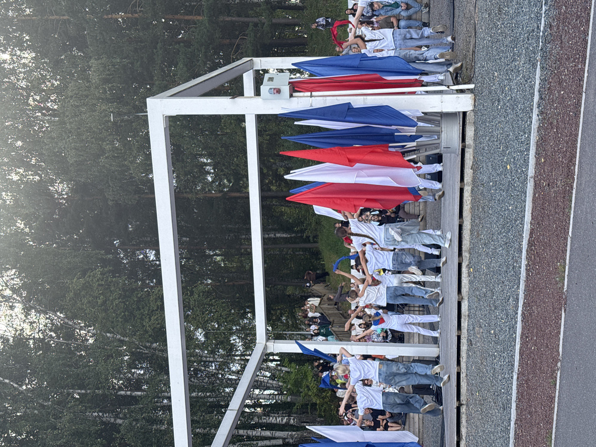 In Chaikovsky, Russia, a group of young people in white t-shirts and jeans are performing on a wooden outdoor stage. They are arranged in a formation and appear to be dancing, with their arms outstretched. Several large flags, predominantly red, white, and blue, are held by individuals on the stage and are also displayed on flagpoles. Behind the stage, an audience is seated on bleachers, watching the performance. The backdrop consists of lush green trees, indicating an outdoor setting surrounded by nature. The overall scene suggests a cultural event or celebration taking place in a park or open-air venue in Chaikovsky.