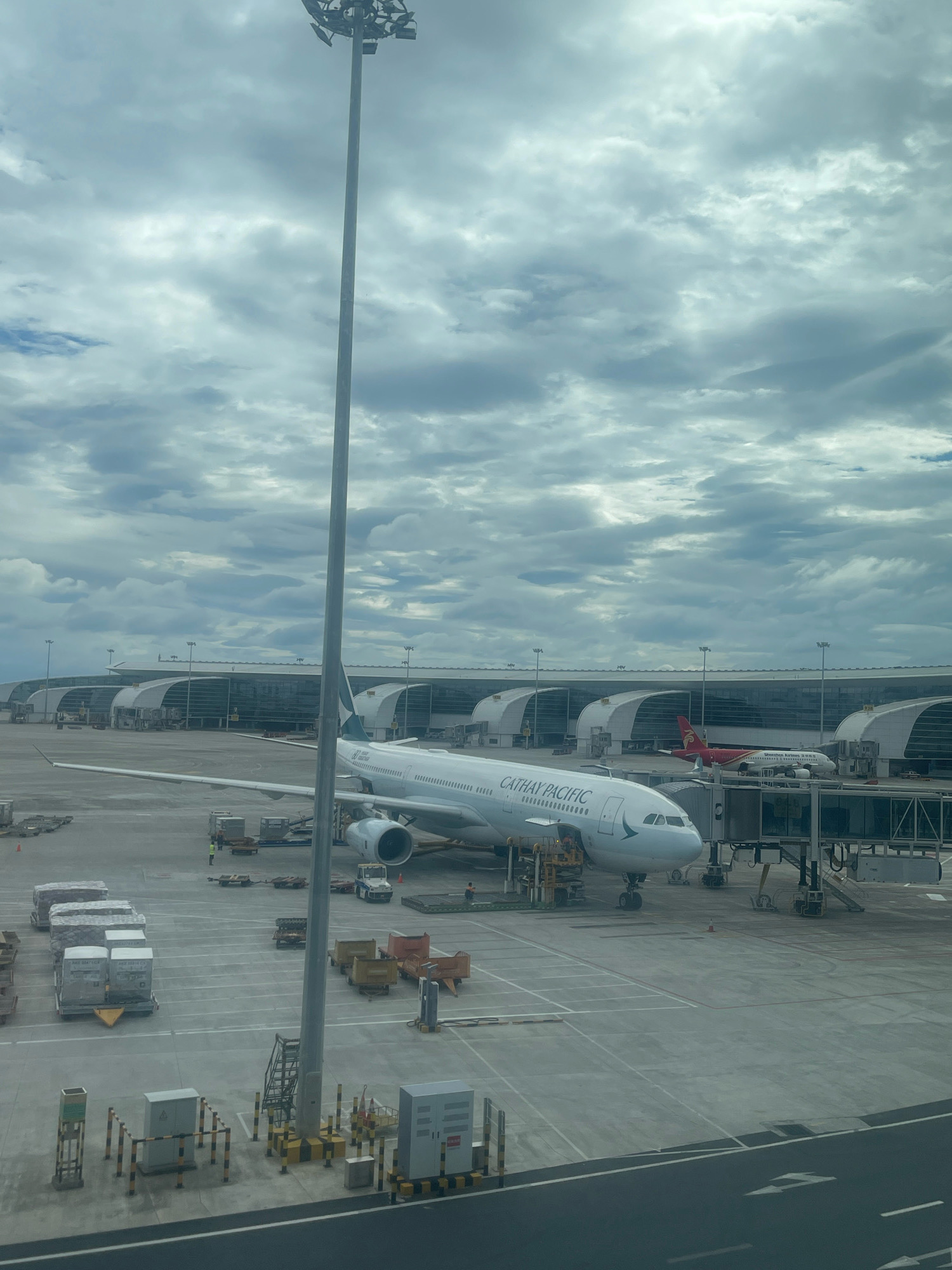 The image shows an aerial view of Guangzhou Baiyun International Airport in Baiyun District, China, on a cloudy day. In the foreground, a Cathay Pacific airplane is parked at a gate, connected to the terminal by a jet bridge. Various airport equipment and baggage carts are visible on the tarmac. In the background, the modern architecture of the airport terminal stretches across the frame, with other aircraft parked at distant gates. A tall light pole is prominently positioned in the left foreground. The sky is filled with a dramatic display of grey and white clouds, suggesting overcast weather.