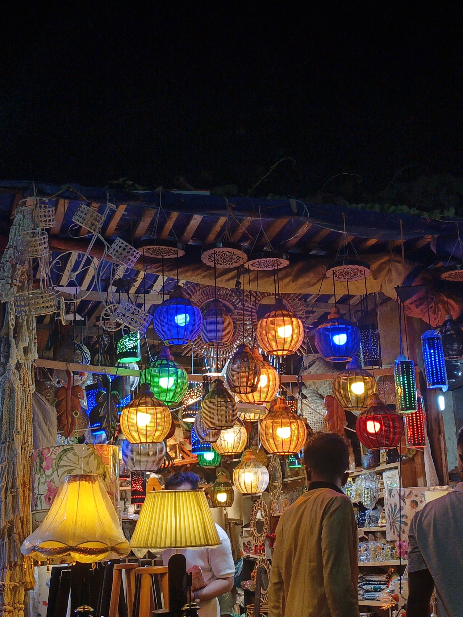 The image depicts a nighttime street market scene in Dhaka, Bangladesh, characterized by a vibrant display of hanging lanterns and decorative items. The foreground features two large, yellow lampshades, one appearing somewhat tattered. Behind these, a diverse array of lanterns are suspended, varying in shape, size, and color, including bright blue, green, orange, and red hues. Some lanterns have intricate woven or perforated designs, allowing the internal light to create patterns.
In the background, a stall is visible with shelves stocked with various goods, including what appear to be small glass figurines, decorative mirrors with ornate frames, and textiles. Two individuals are partially visible in the scene; one person, viewed from behind and in profile, is wearing a light brown or yellow long-sleeved shirt and is looking towards the merchandise. Another person, also seen from behind, is wearing a white shirt and is positioned to the right of the frame. The market stall itself is constructed with wooden elements and covered with striped blue and white fabric. The overall atmosphere suggests a bustling evening market, likely a tourist-oriented or popular local shopping area within Dhaka.