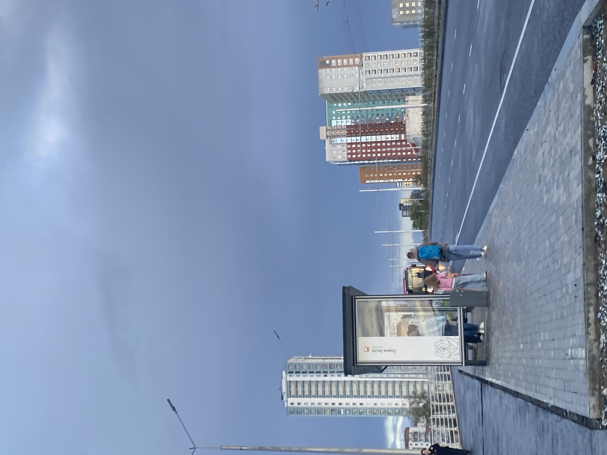 This image shows a bus stop in Perm, Russia, on a cloudy day. In the foreground, a paved sidewalk leads to a bus stop shelter with a poster inside. Three people are standing near the shelter: a woman in a pink shirt and jeans, a man with a blue backpack, and another person partially obscured. A bus is visible behind the shelter. In the background, several tall residential buildings with colorful facades are visible, as well as a street lamp and overhead wires. The sky is dark and overcast, suggesting impending rain.