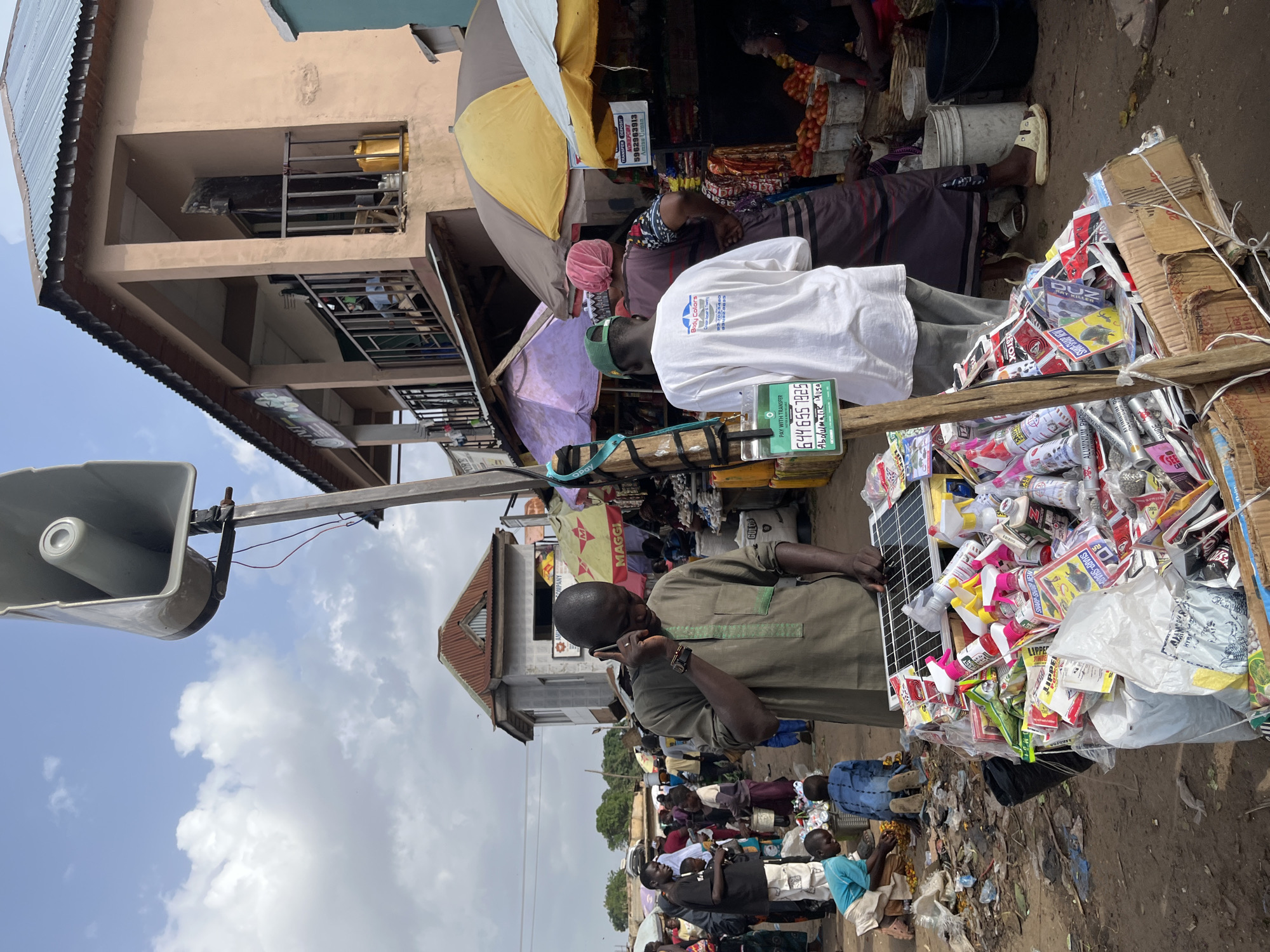 The image depicts a busy street market in Rafin Par, Nigeria. In the foreground, a man in a green tunic is talking on a mobile phone while standing beside a stall piled high with various goods, including solar panels, spray cans, and packaged items. A wooden pole with a large speaker is positioned to the left of the man. Behind him, another person in a white t-shirt and green cap is visible, along with several market stalls covered by umbrellas, displaying goods such as tomatoes. People are milling about the market, suggesting active commerce. The environment appears somewhat cluttered, with debris on the ground, and buildings with corrugated metal roofs are visible in the background. The sky is partly cloudy.