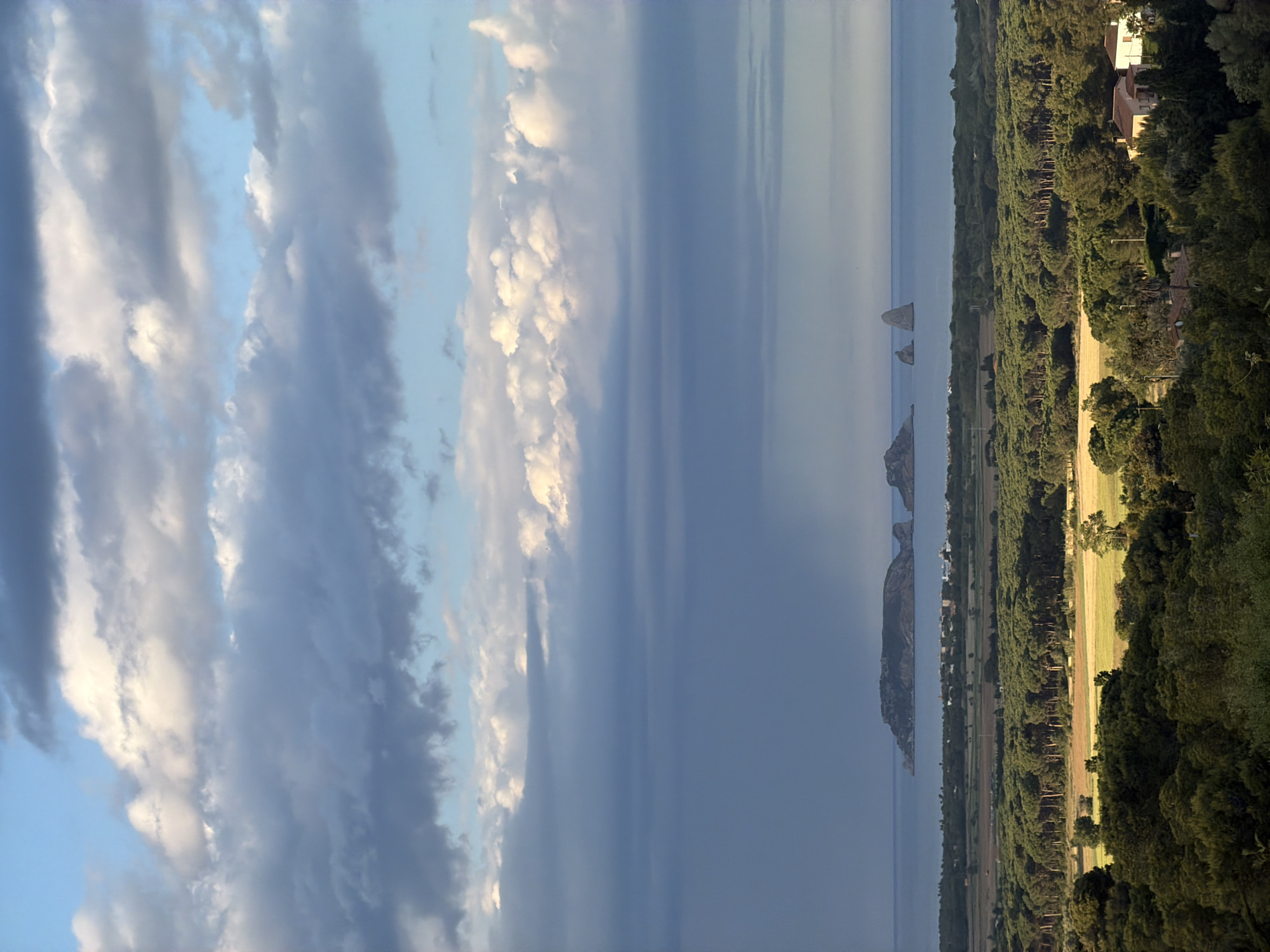 The image shows a view from the city of Pals, Spain, possibly looking eastward over the coastline. The sky occupies a large portion of the left side of the frame, with striated, layered clouds and patches of blue. A body of water stretches across the center. In the distance, rock formations jut out of the water. On the right is a forested area with scattered buildings, mostly obscured by trees. Some fields are visible among the trees.
