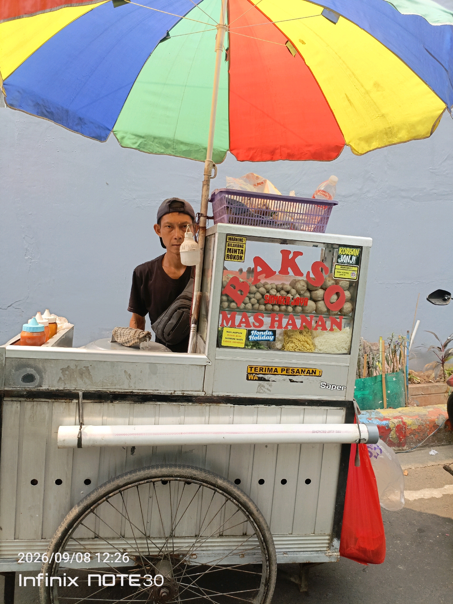 A street vendor, a man of Southeast Asian descent with a dark baseball cap and black t-shirt, sits behind a mobile food cart under a large, multi-colored umbrella. The cart is made of corrugated metal and displays a sign advertising "BAKSO SUMBER JAYA MAS HANAN," with meatballs and noodles visible inside a glass display case. To the left of the vendor are condiment bottles, and to the right, a red plastic bag hangs from the cart. The background is a plain blue wall. The scene appears to be in a city, possibly Central Jakarta, Indonesia, given the context of a "bakso" cart and the provided timestamp and phone model.
