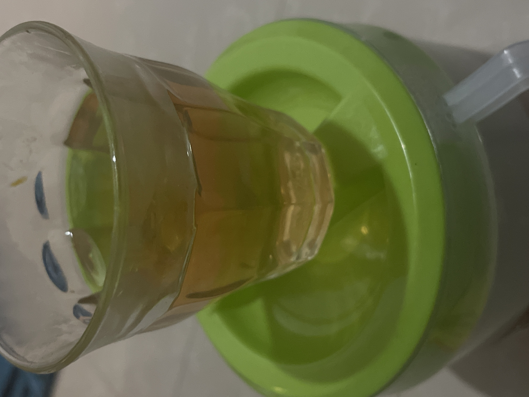 A clear glass containing a golden-amber liquid sits atop a bright green plastic container. The glass has a subtly ribbed texture. The liquid fills about two-thirds of the glass. The green container has a smooth, slightly curved surface. A light gray, rectangular handle or spout protrudes from the right side of the green container. The background is a neutral, light-colored surface, possibly a countertop or table. The lighting appears to be artificial and slightly diffused, casting subtle shadows. No people are visible in the image.