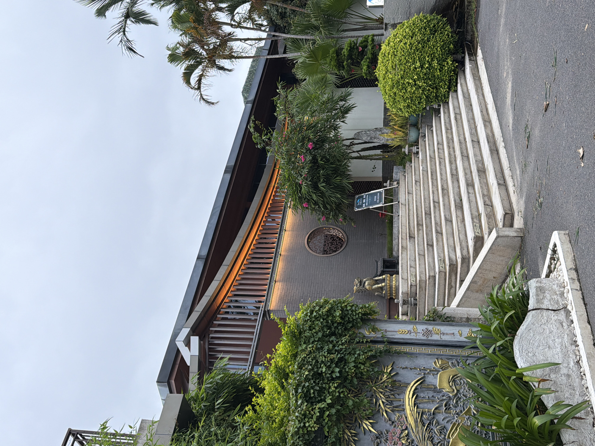 The image shows the exterior of a building with a modern design incorporating traditional Chinese architectural elements, located in Longgang District, China. A wide, multi-tiered staircase made of stone leads up to the entrance of the building. The building itself has a dark-colored, sloped roof with wooden accents and integrated orange lighting. The facade features grey brickwork and a decorative circular window with intricate metalwork. Lush green vegetation, including trees, bushes, and vines, surrounds the building and the staircase, with a large, ornate golden metal gate to the left featuring bamboo and floral motifs. A golden lion statue sits on the second step of the staircase, next to a small decorative pagoda. A sign on an easel stands near the middle of the stairs. Palm trees and other tropical plants are visible in the background. The sky is overcast. There are no people visible in the image.