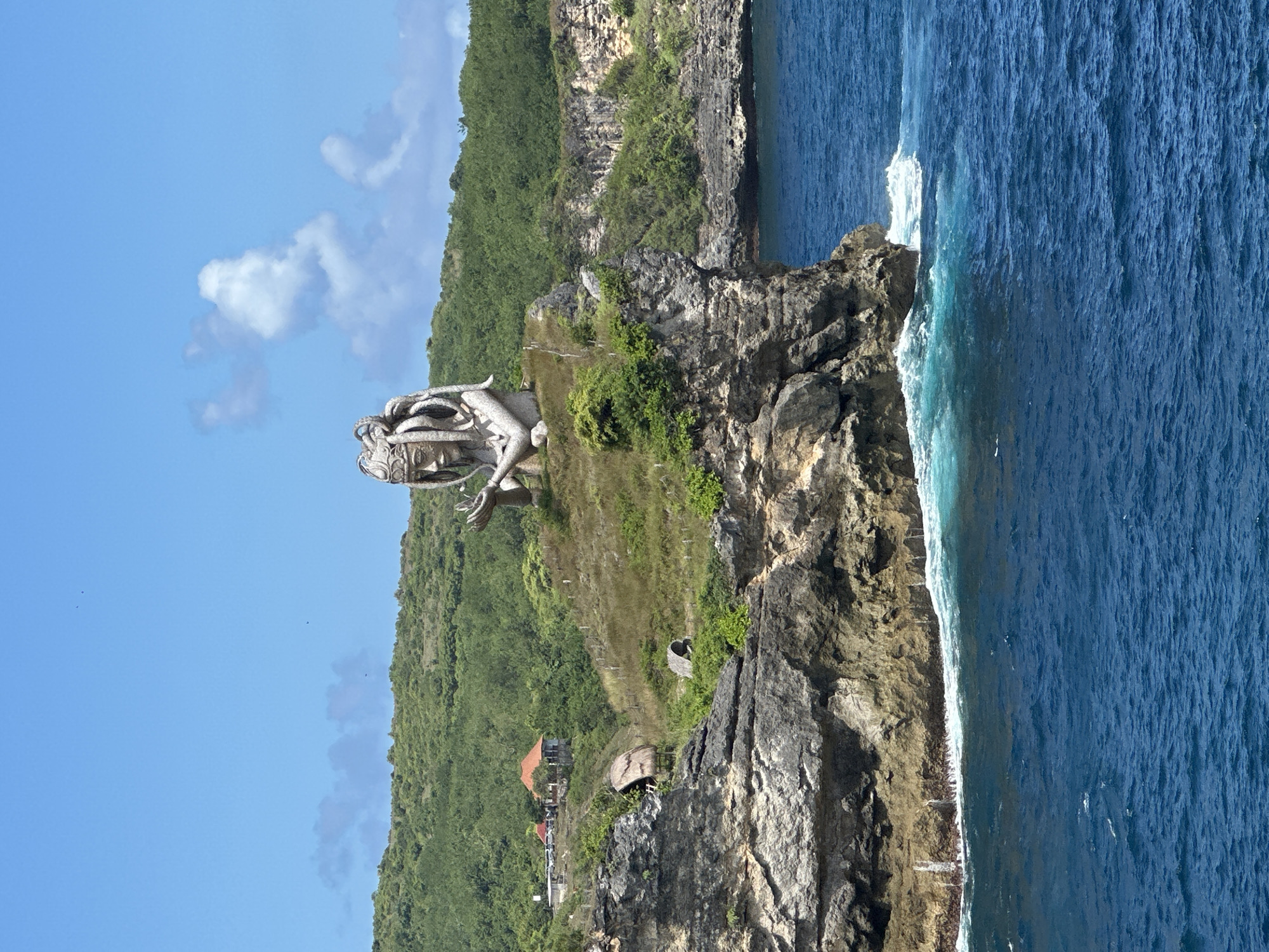 The image depicts a coastal scene in Nusa Penida, Indonesia, featuring a large statue on a rocky outcrop. The statue, rendered in a light-colored material, is positioned prominently on the cliff edge overlooking the ocean. The coastline is characterized by rugged cliffs and lush green vegetation, which covers the terrain leading up to the cliffs. A small structure is visible at the base of the cliff. The ocean stretches out towards the horizon, its surface a deep blue. The sky is blue with scattered clouds. The overall impression is of a picturesque coastal landscape with a cultural landmark.