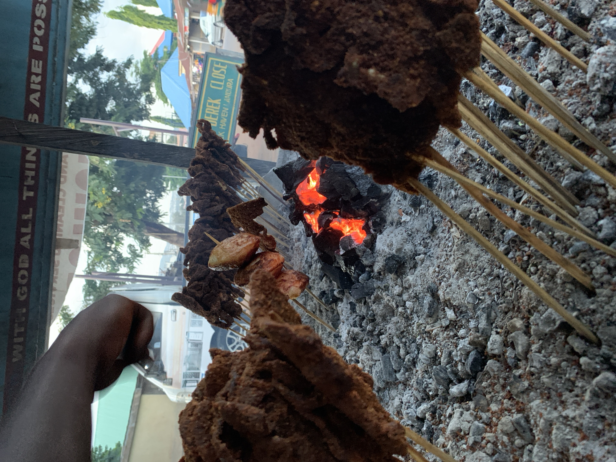 This media file captures a vibrant outdoor street food scene, likely centered around grilling. The primary subjects are numerous skewers of dark, seasoned meat, characteristic of Suya, a popular West African grilled meat. These skewers are positioned over an active charcoal grill, with bright red embers visible at its heart, indicating intense heat for cooking. Surrounding the embers are ashes and small pieces of charcoal, suggesting prolonged use of the grill.
A dark-skinned hand is partially visible on the left side of the frame, appearing to tend to the skewers or the grilling process. The meat on the skewers varies slightly, with some pieces appearing more solid and coated, while others show some lighter, possibly fattier, parts.
In the background, various elements suggest an urban or semi-urban outdoor setting during daylight. Green foliage from trees is visible, along with parts of buildings and what appears to be a light-colored vehicle. There are also hints of colorful awnings or structures.
Notable text includes a sign that reads "DEREK CLOSE" and "K HAVEN / ANDURA," possibly indicating a street name or a business location. Another partial sign displays the words "WITH GOD ALL THING S ARE POSS," likely part of the phrase "With God all things are possible." These signs and the type of food align with the provided context of Jos, Nigeria, painting a picture of a bustling local food vendor. The overall impression is one of active food preparation in a public space.