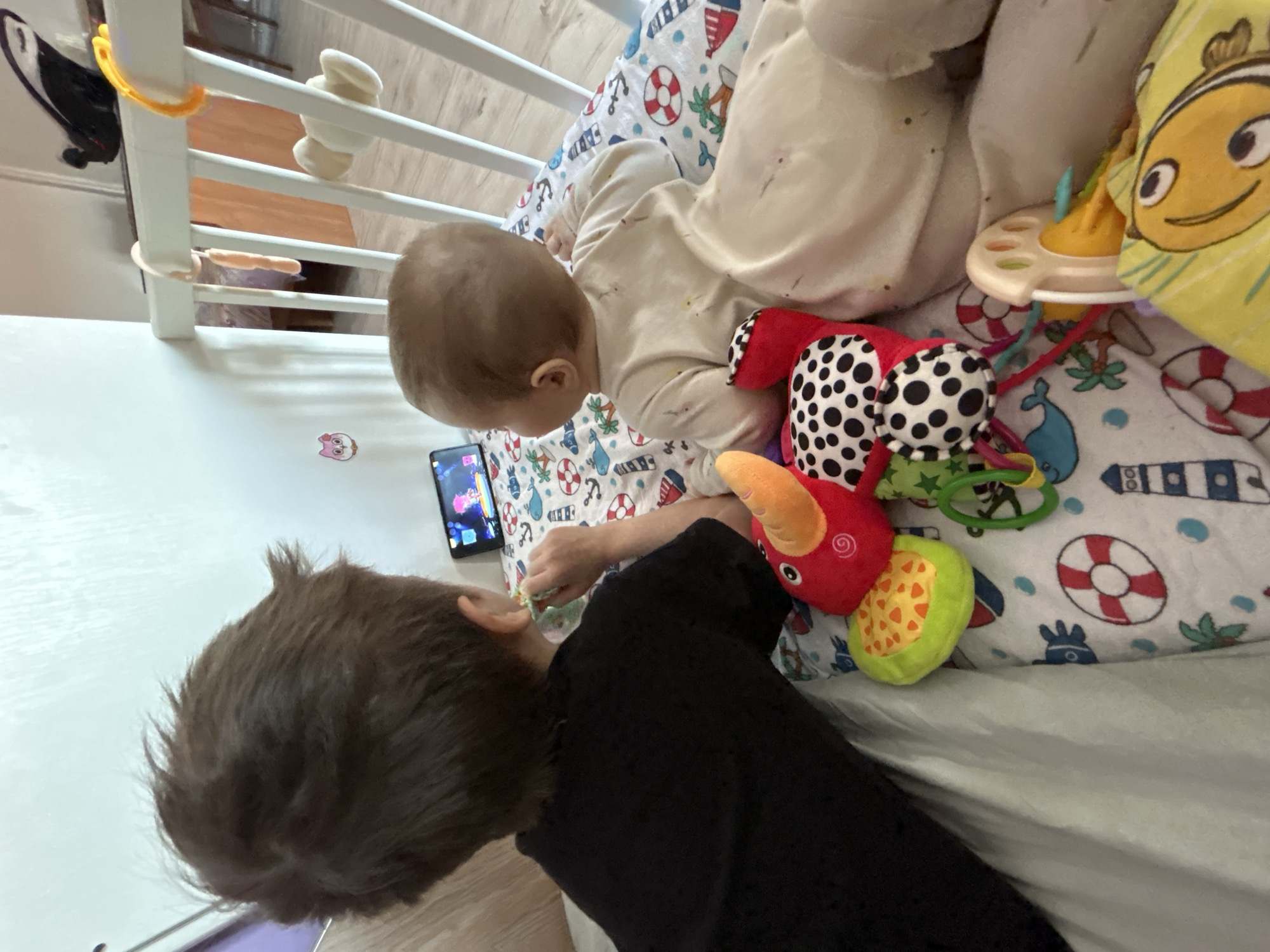 A top-down shot depicts an indoor scene. A baby in light-colored pajamas lies in a crib with nautical-themed bedding. A colorful soft toy is near the baby. An older child, with dark hair and wearing a black shirt, leans into the crib, possibly interacting with the baby. A smartphone is visible on the crib mattress. The location is Rusalevka, Russia.