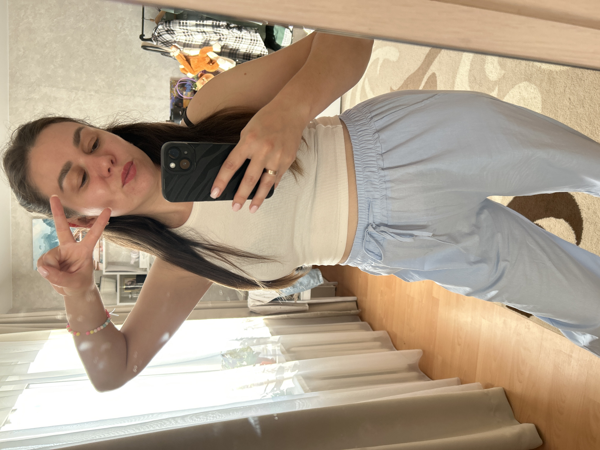 A woman is taking a mirror selfie in what appears to be a bedroom. She is wearing a white tank top and light blue pants. She is holding a phone in her right hand and making a peace sign with her left hand. The background includes a window with sheer curtains, a dresser with clothing on it, and a shelf with various items. The floor is wooden, and there is a rug with a patterned design. The overall impression is a casual, indoor setting. No specific location in the city of Фоки, Russia can be determined from the visual information.