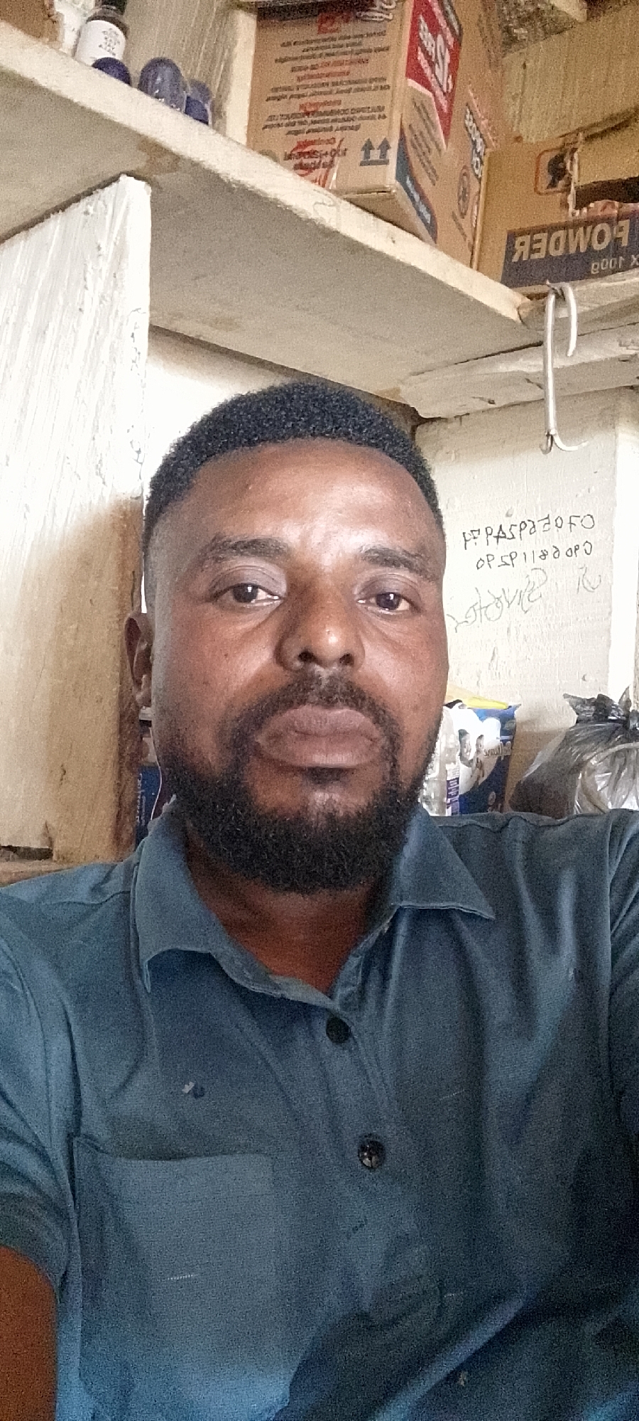 A man with dark, short, curly hair and a beard is pictured in a close-up, waist-up shot. He is wearing a blue collared shirt with two buttons visible. The background appears to be an interior space with shelves containing various items, including boxes and bags. There is writing on the wall behind him, some of which is illegible. The lighting suggests it might be indoors with artificial light. The image does not contain information about the city of Durimi, Nigeria or any specific location within it.