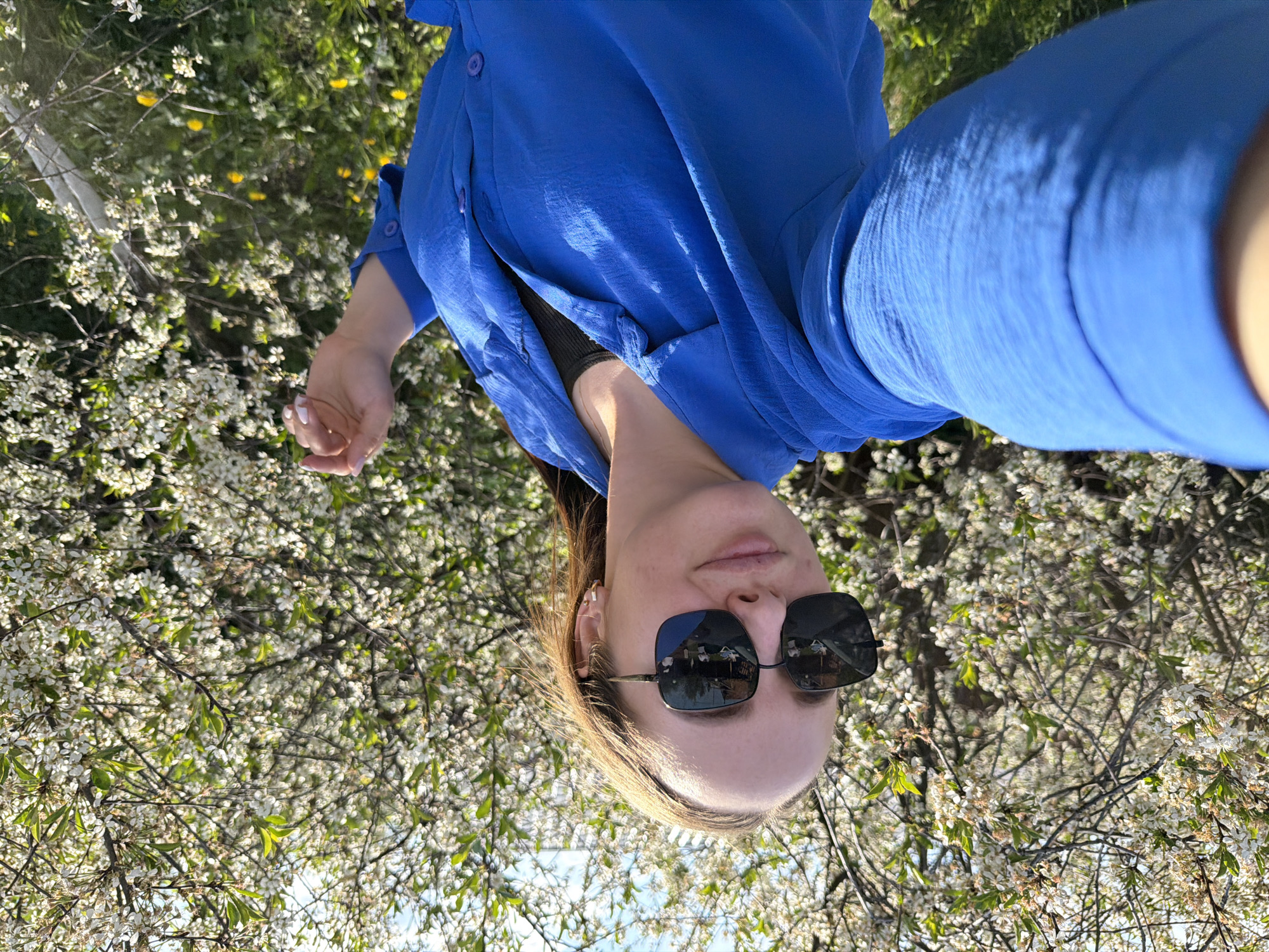 A selfie taken outdoors in Chaikovsky, Russia, shows a woman wearing sunglasses and a blue button-down shirt. The background is filled with blooming trees with small white flowers and green foliage. The woman is positioned close to the camera, with her face centered in the lower half of the frame. The image has been rotated counter-clockwise.