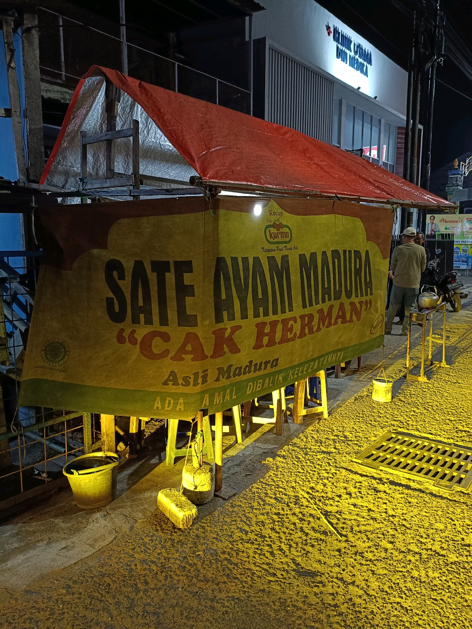 The image depicts a street-side food stall at night in Purwomartani, Indonesia. The stall features a large yellow banner with dark text that reads "SATE AYAM MADURA 'CAK HERMAN' Asli Madura". Below this, smaller text says "ADA AMAL DIBALIK KELEZATANNYA". The stall is covered by a red tarp, supported by a wooden frame. Several yellow chairs are visible underneath the stall. A yellow bucket and a rectangular object are placed on the ground in front of the stall. The ground is paved and illuminated with a yellow light. In the background, a building with "KLINIK UTAMA DWI MEDIKA" is visible. A motorcycle is parked to the right of the stall, and two individuals are standing near it. The overall scene suggests a typical roadside food vendor operating in a populated urban area during the evening.