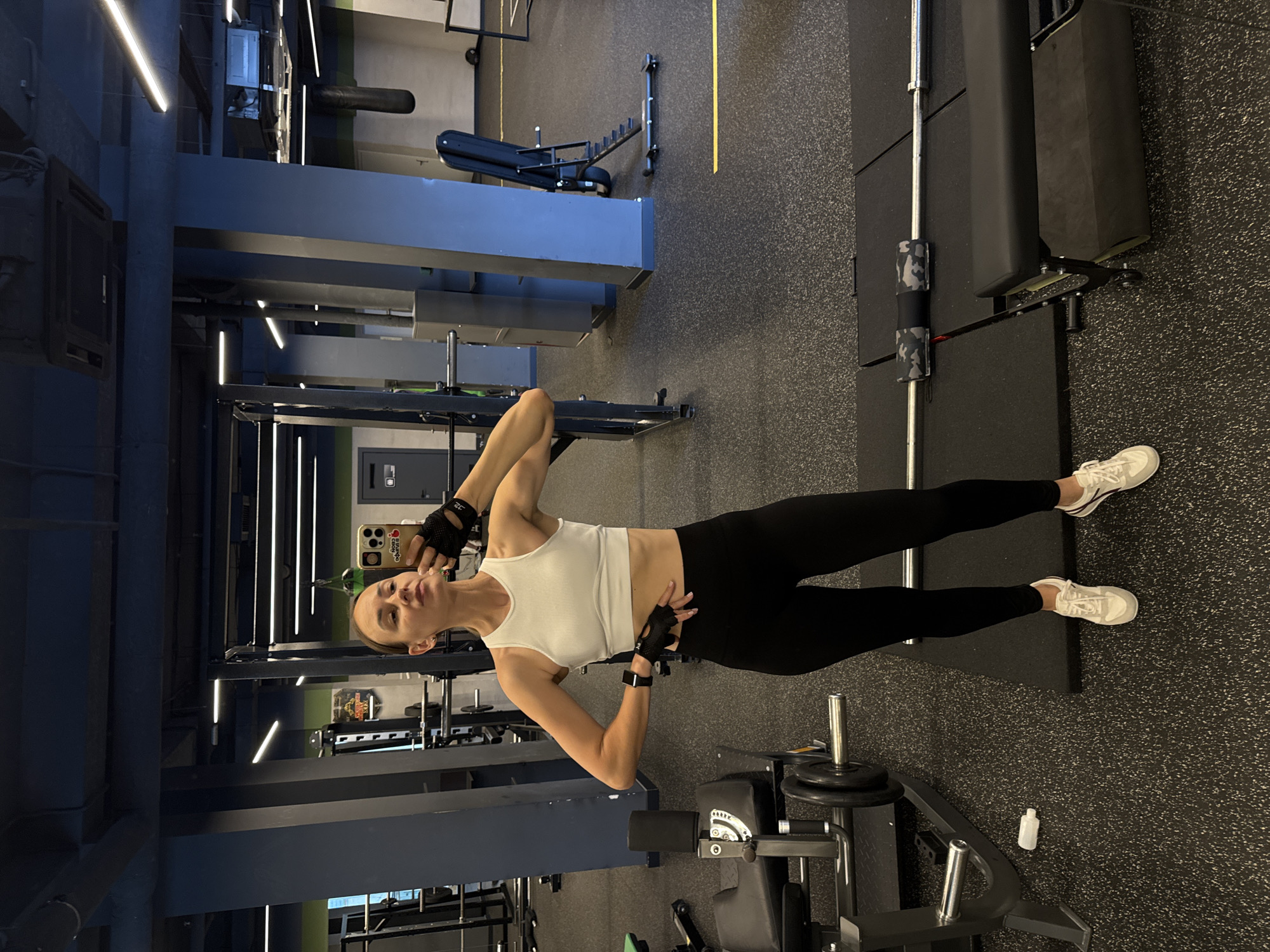 A woman is taking a mirror selfie in a gym. She is wearing a white crop top, black leggings, black weightlifting gloves, and white athletic shoes. She has her hair pulled back in a bun. She is standing in front of a squat rack. In the background, there are various gym equipment, including weight machines, a bench press, and a punching bag. The gym appears to be well-lit and spacious. The woman is posing with her left hand on her hip and her right hand holding her phone. The image is taken from a full-body perspective, showing her from head to toe. The overall impression is of a woman who is fit and confident, enjoying her workout.