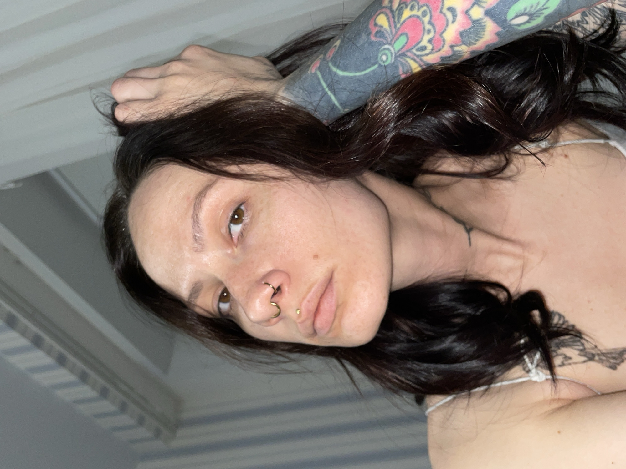 A woman with dark hair and fair skin is shown in a close-up, angled shot. She has a nose ring and a labret piercing. Her right arm, which is heavily tattooed with floral and geometric designs, is raised near her head. She is wearing a light-colored top with thin straps. In the background, a portion of a white ceiling is visible. The location is not evident from the image.