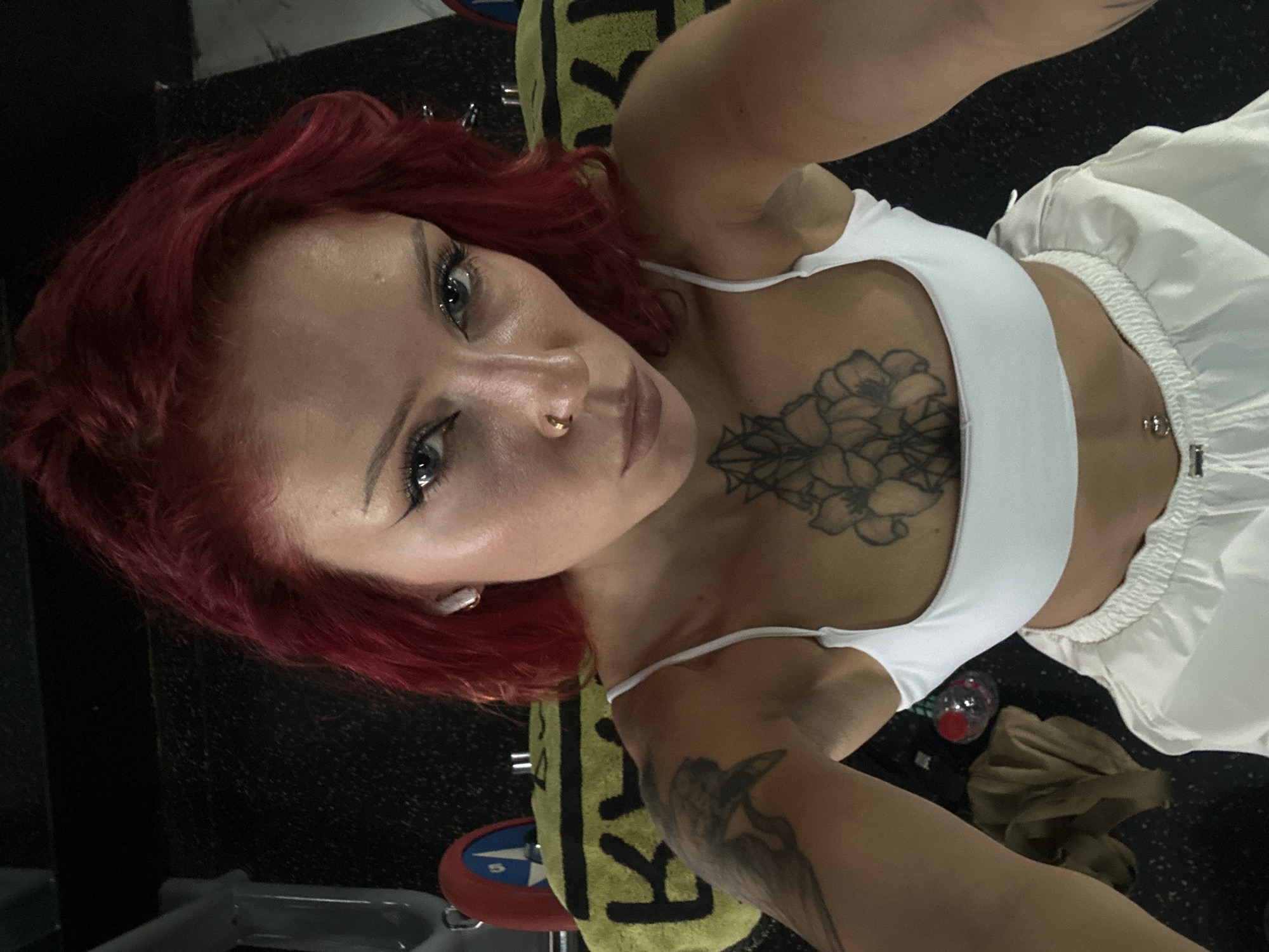A woman with red hair, wearing a white sports bra and white pants, takes a selfie. She has a floral tattoo on her chest. The environment appears to be a gym. There is a towel with black lettering and a weight plate visible. The location is North Kuta, Indonesia.