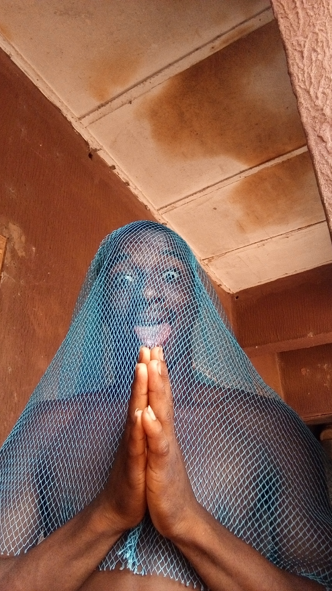 An individual with dark skin tone is seen indoors, partially obscured by a light blue net. The hands are clasped together and positioned in front of the face. The interior is painted in earth tones, and there are stained ceiling tiles. The image does not show sufficient information to determine that the location is Onitsha, Nigeria.