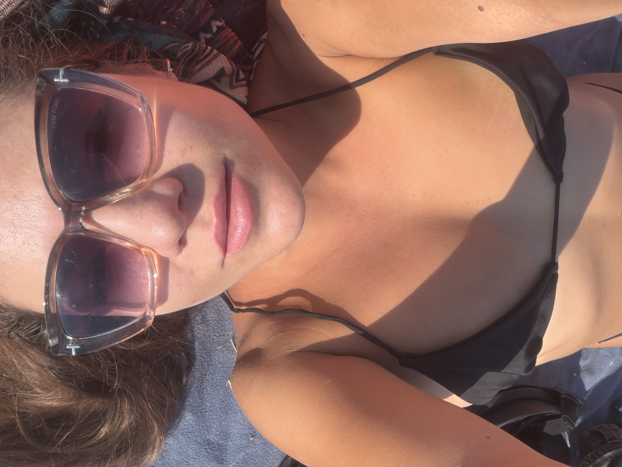 The image shows a close-up, overhead view of a person lying down, wearing a black bikini top and sunglasses. The person's face is partially visible, with their lips painted a soft pink and wearing large, light-brown tinted sunglasses with "TOM FORD" written on the arm. Sunlight casts strong shadows across their chest and shoulder. Their hair is dark brown and appears slightly disheveled, with a patterned scarf visible behind their head. The setting appears to be outdoors, possibly at a beach or poolside, with a blue towel or blanket underneath. The lighting suggests it is daytime, with the sun high in the sky. The overall impression is one of relaxation and enjoying a sunny day.