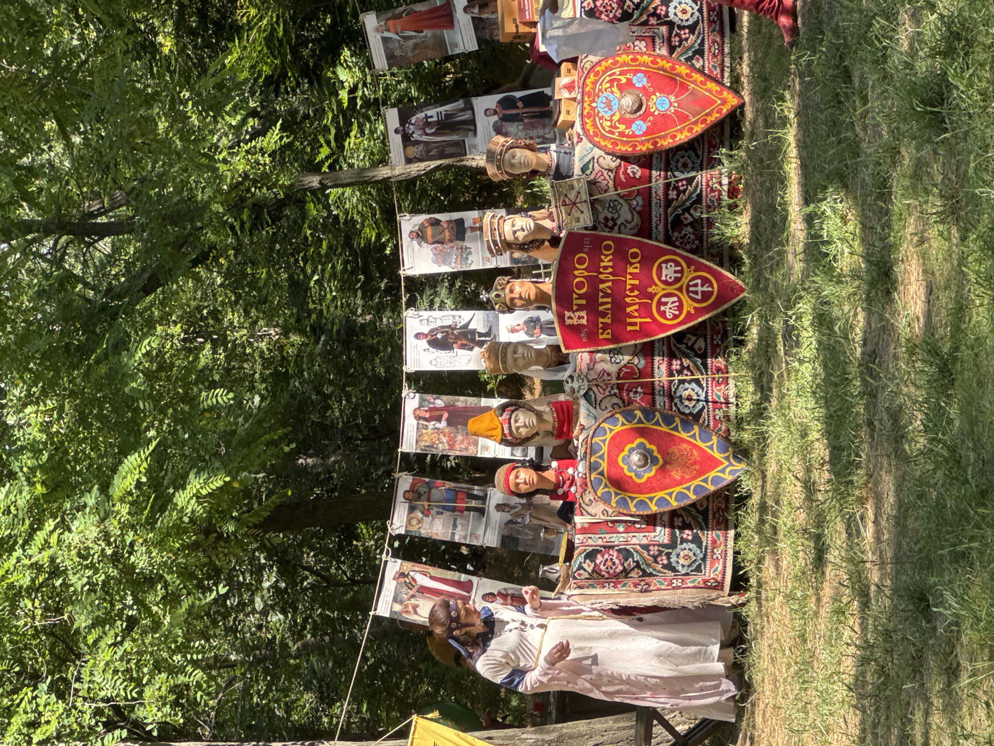 The image captures an outdoor historical reenactment or exhibition. In the foreground, a table is draped with vibrantly patterned textiles, displaying several mannequins dressed in historical attire and wearing crowns or elaborate headwear. Two shields, red with intricate yellow and blue designs, are prominently placed in front of the table, one featuring Cyrillic script that reads "ВТОРО БЪЛГАРСКО ЦАРСТВО" (Second Bulgarian Empire) and what appears to be a cross symbol. Behind the table, a woman in a long, pale pink robe with blue trim stands to the left, looking down at the table. A string of posters hangs behind the setup, depicting historical figures in various medieval clothing. The backdrop consists of lush green trees, indicating an outdoor setting, likely within a park or natural area. The ground is covered with grass and dappled sunlight. Based on the signage, this event is likely taking place in Plovdiv, Bulgaria, given the reference to the Second Bulgarian Empire, a significant historical period for the region.