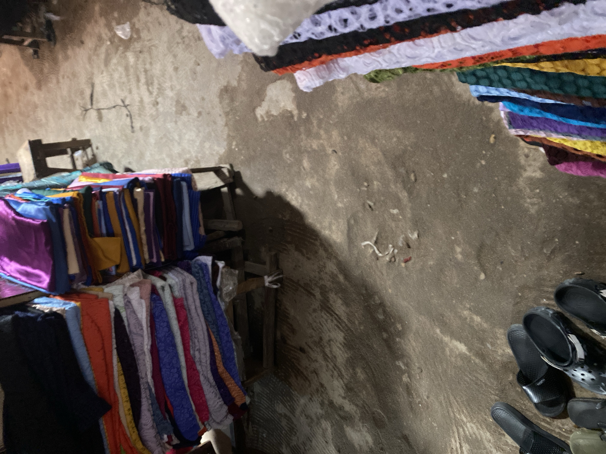 This image captures an outdoor market scene in Jalingo, Nigeria. The primary subjects are an assortment of fabrics and clothing items displayed for sale, along with several pairs of black footwear, possibly for sale or belonging to a vendor. The scene is set on a rough, concrete ground, likely a marketplace stall.
The fabrics are arranged in stacks, with some appearing to be colorful, patterned textiles and others simpler, solid-colored materials like satin. There are also rolls of what looks like lace or trim in black, white, and orange colors. The clothing items include what seem to be dresses or skirts, displayed on hangers or draped. The footwear consists of various styles of black sandals and flip-flops.
The lighting suggests it might be daytime, but the quality of light is not distinct enough to determine the exact time. There are no clear indications of weather conditions. The rough ground and the way the goods are displayed are typical of an informal market setting.
There is no visible text in the image. The overall impression is a bustling, everyday market environment where various goods are being offered. The focus is on the colorful array of fabrics and the practical items like footwear.
