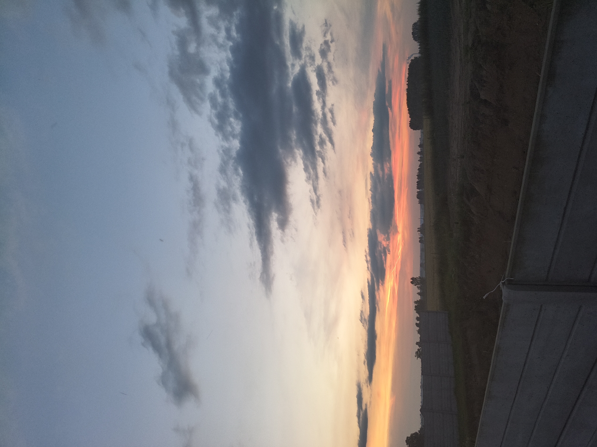 This image depicts a rural landscape at dusk, likely in or near Galonki, Poland. The sky transitions from a pale blue at the top to vibrant hues of orange and pink near the horizon, indicating a sunset. Dark, scattered clouds drift across the sky. In the foreground, a concrete fence, constructed from modular panels, runs horizontally across the frame, suggesting an area under construction or a boundary. Beyond the fence, a field is visible, with darker areas of soil and lighter patches that could be vegetation or tilled land. A line of trees forms a distant treeline, and a few structures, possibly buildings or agricultural elements, are faintly discernible in the far distance. The overall scene is serene and devoid of human presence.