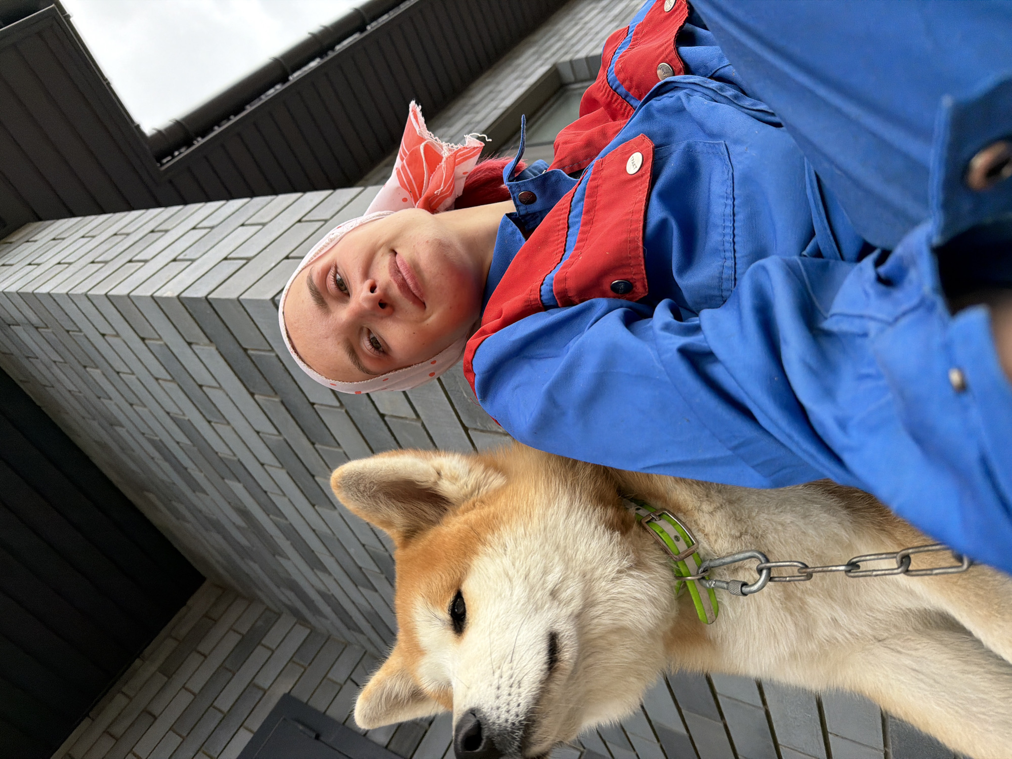 An outdoor, low-angle, close-up selfie shows a young woman with red hair and a light pink and red headscarf. She is wearing a blue and red jumpsuit. To her left, an Akita dog with light brown and white fur sits beside her. The dog has a light green collar with a silver chain leash. They are positioned in front of a grey brick wall with dark wooden eaves visible above. The scene is likely in the city of Chaikovsky, Russia.